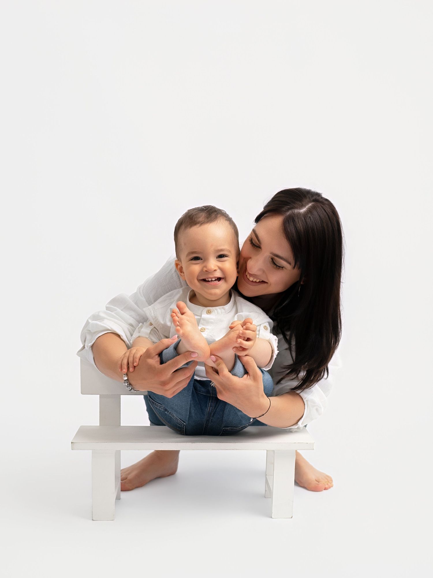 A smiling mother holds her laughing baby on a small white bench against a clean white background.