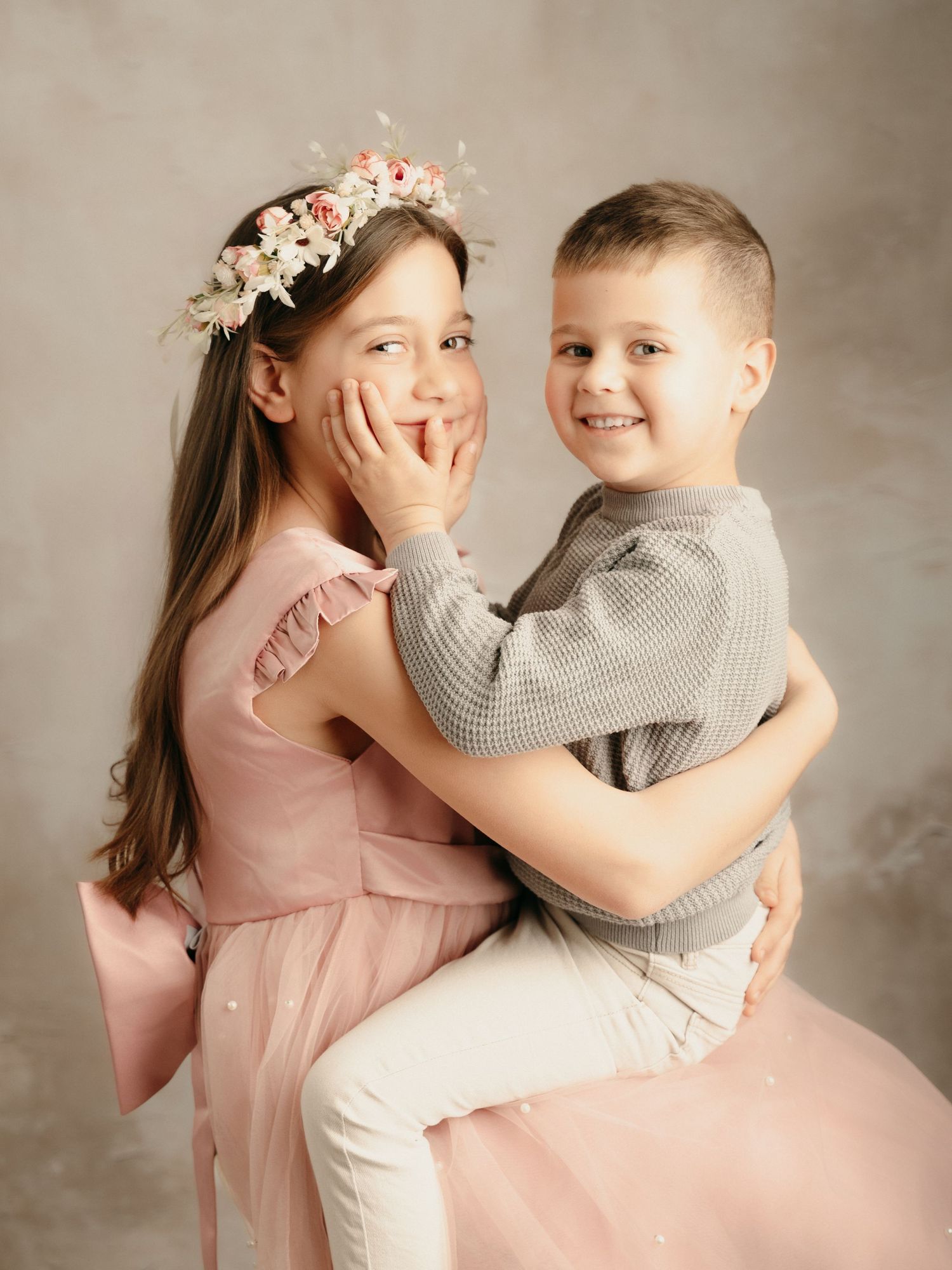 Young girl in pink dress with floral crown hugging her smiling little brother in a studio portrait.