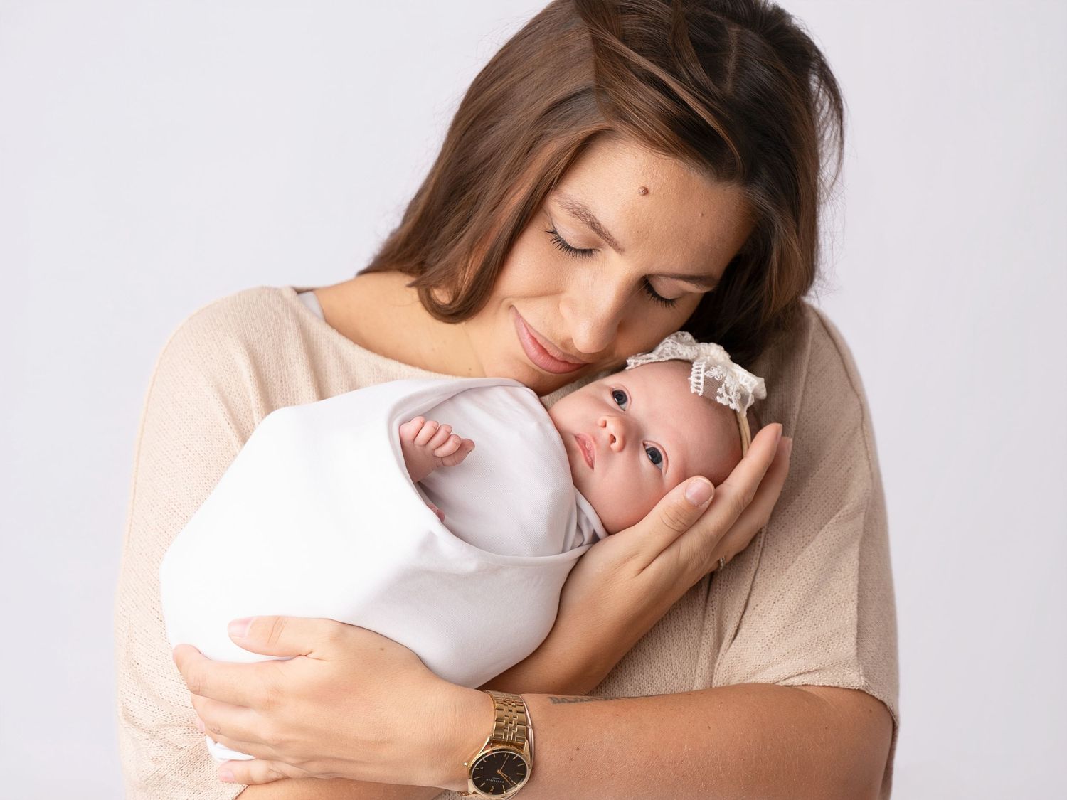 A mother tenderly holds her swaddled newborn baby close, both wearing white, against a soft gray background.