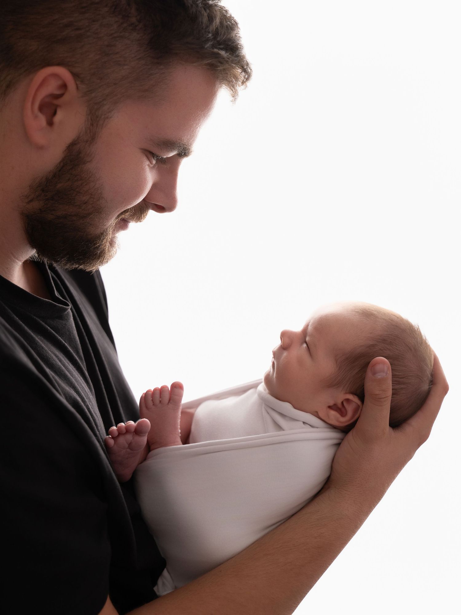 A bearded father tenderly holds his swaddled newborn baby, gazing down lovingly against a white background.