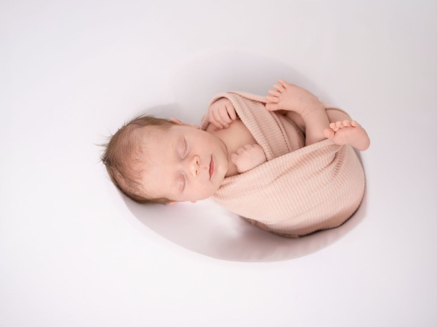 Newborn baby sleeping peacefully wrapped in a soft pink swaddle on a white background.