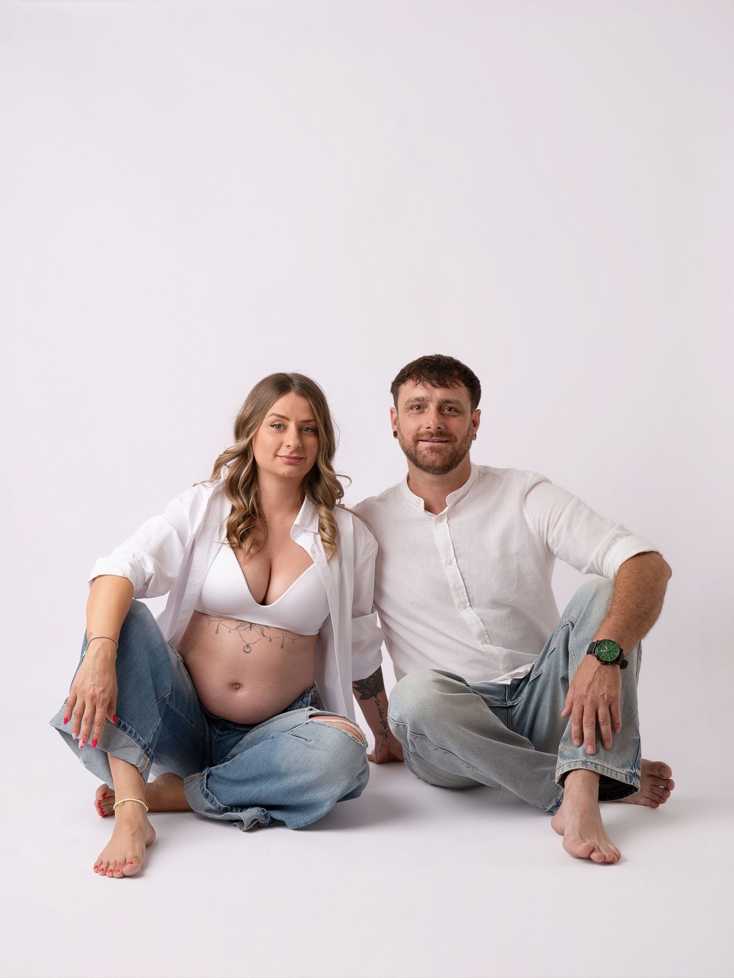 Pregnant couple sitting on floor in white shirts and jeans, smiling at camera in bright studio setting.