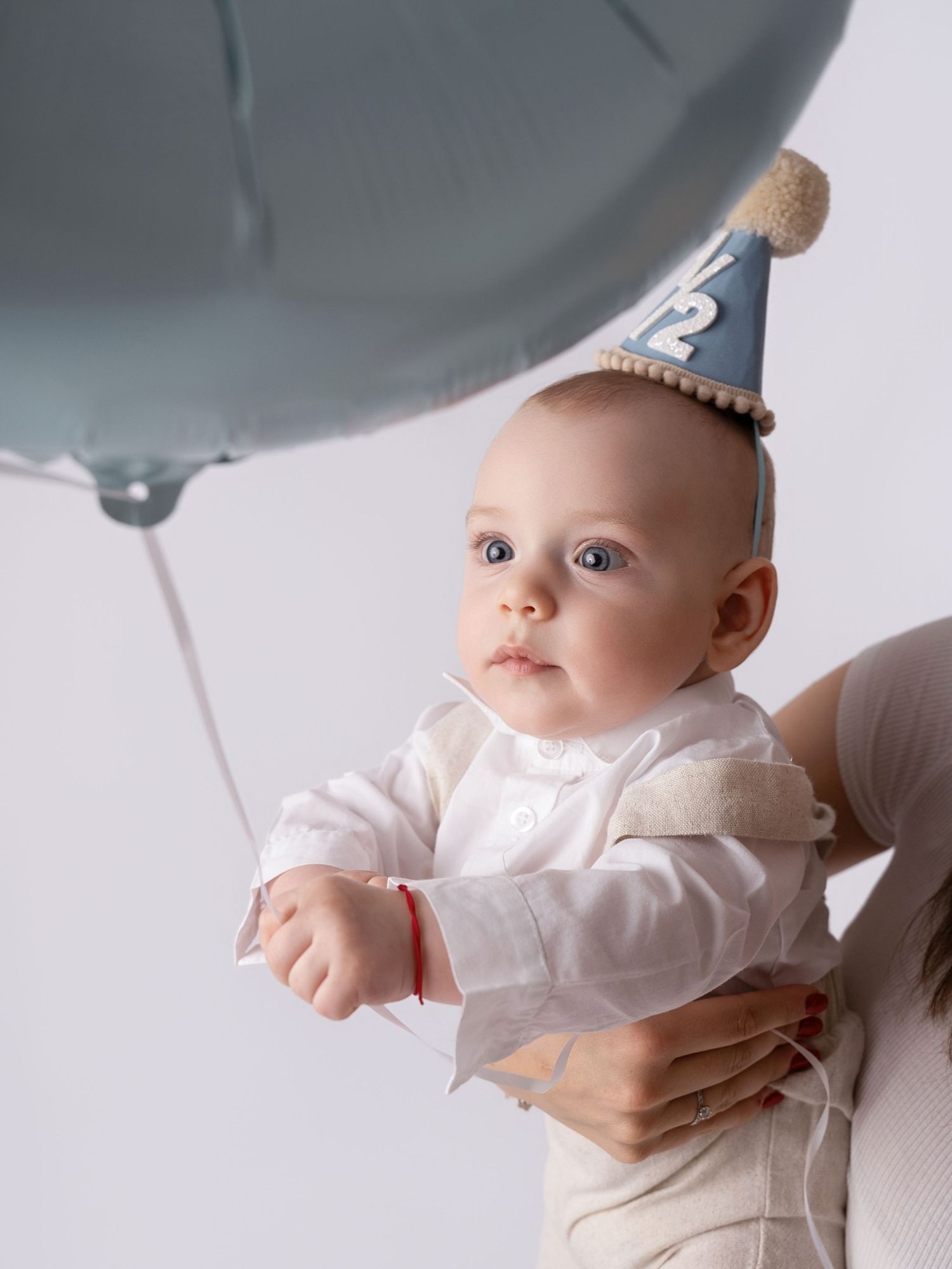 A baby wearing a blue party hat and white outfit holds a gray balloon while being held by an adult.