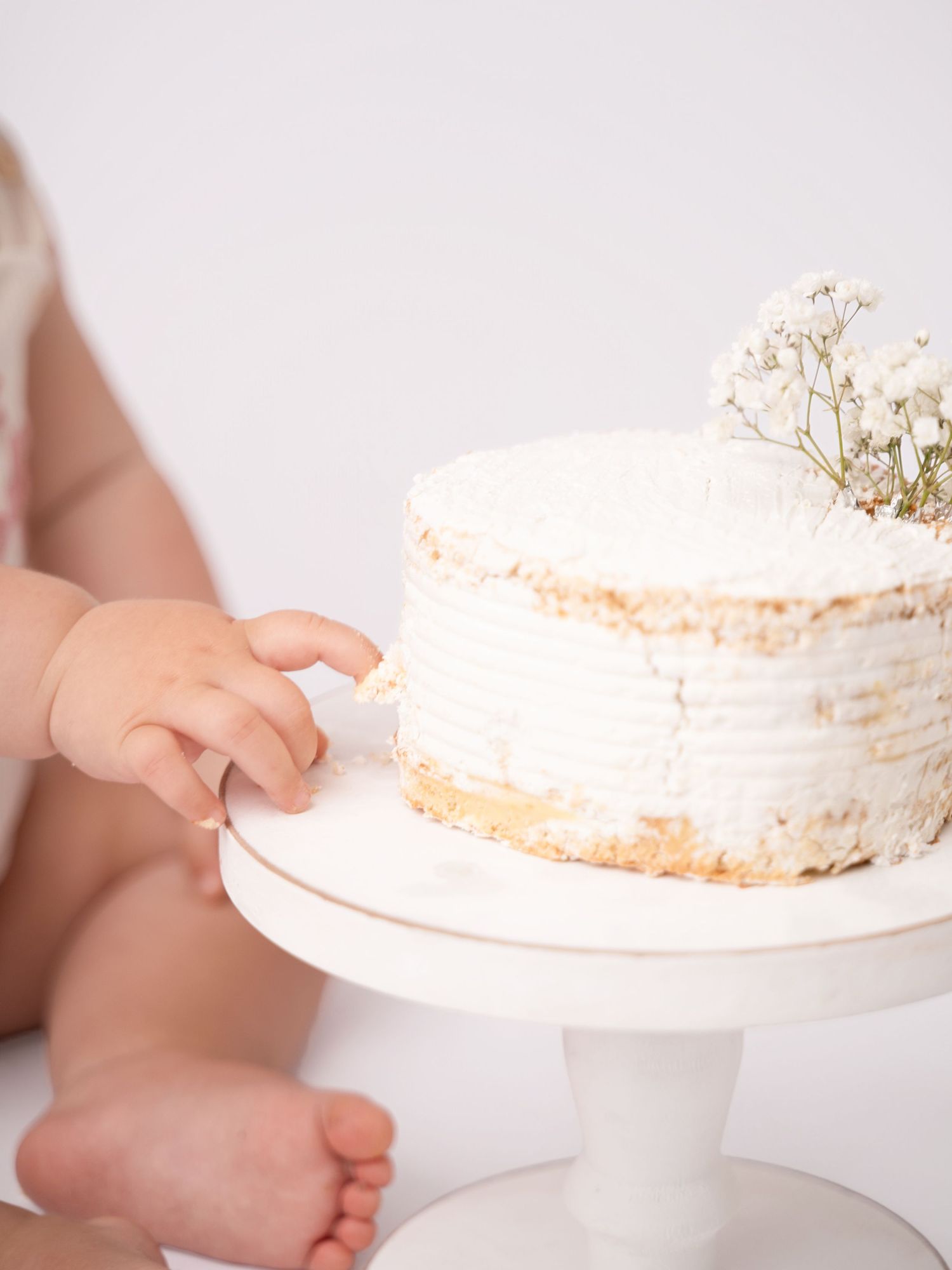 Baby reaching for a white frosted first birthday smash cake topped with baby's breath flowers on a white cake stand.