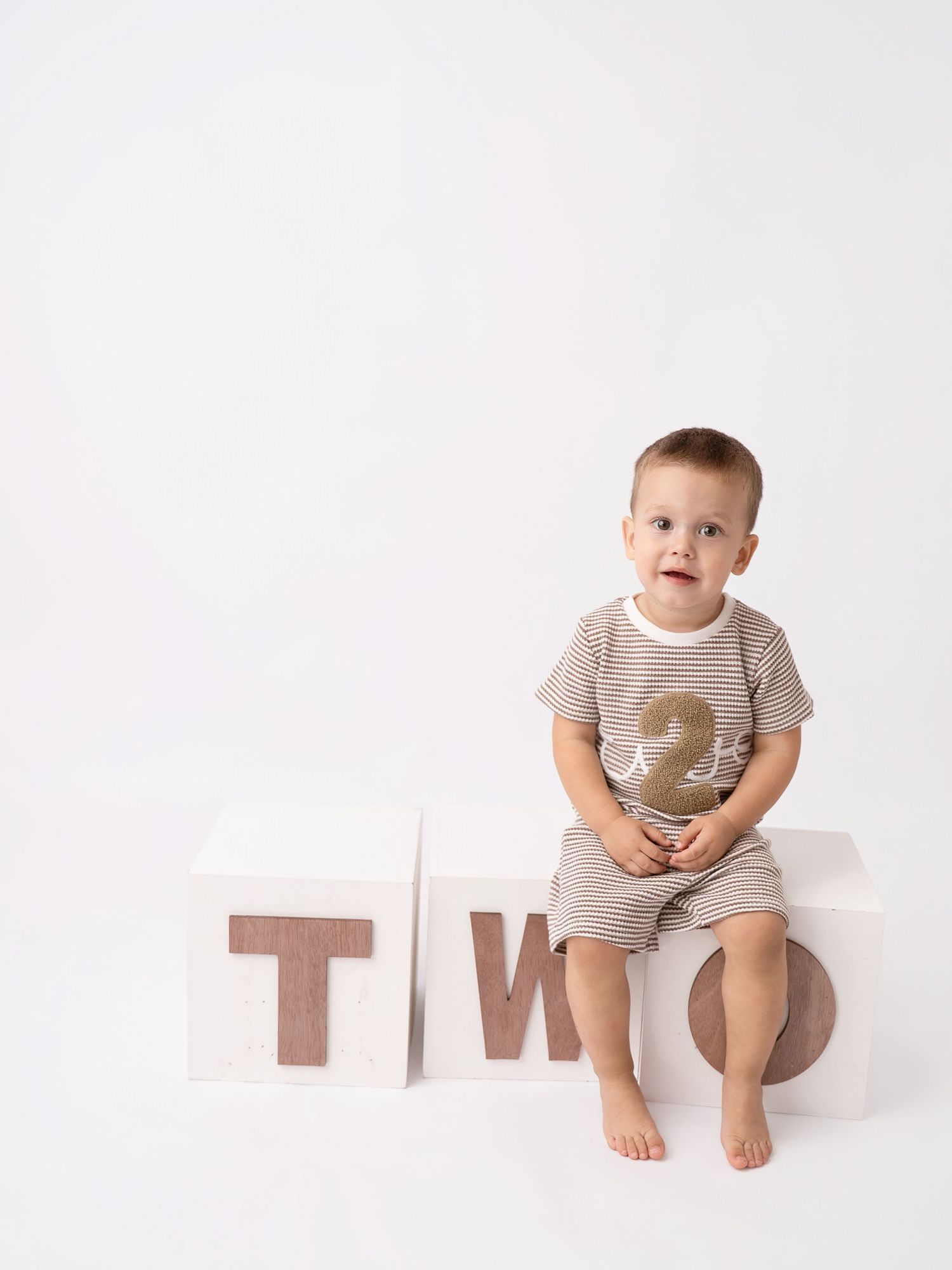 Toddler boy in striped shirt sitting on wooden block spelling TWO for a 2nd birthday photo session.