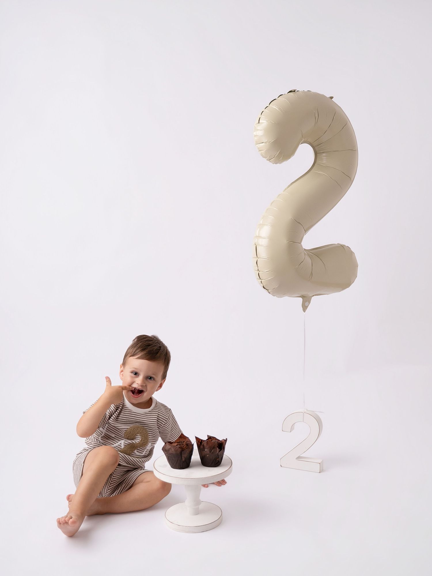 Smiling toddler celebrating 2nd birthday with chocolate cupcakes, number 2 balloon, and white cake stand.