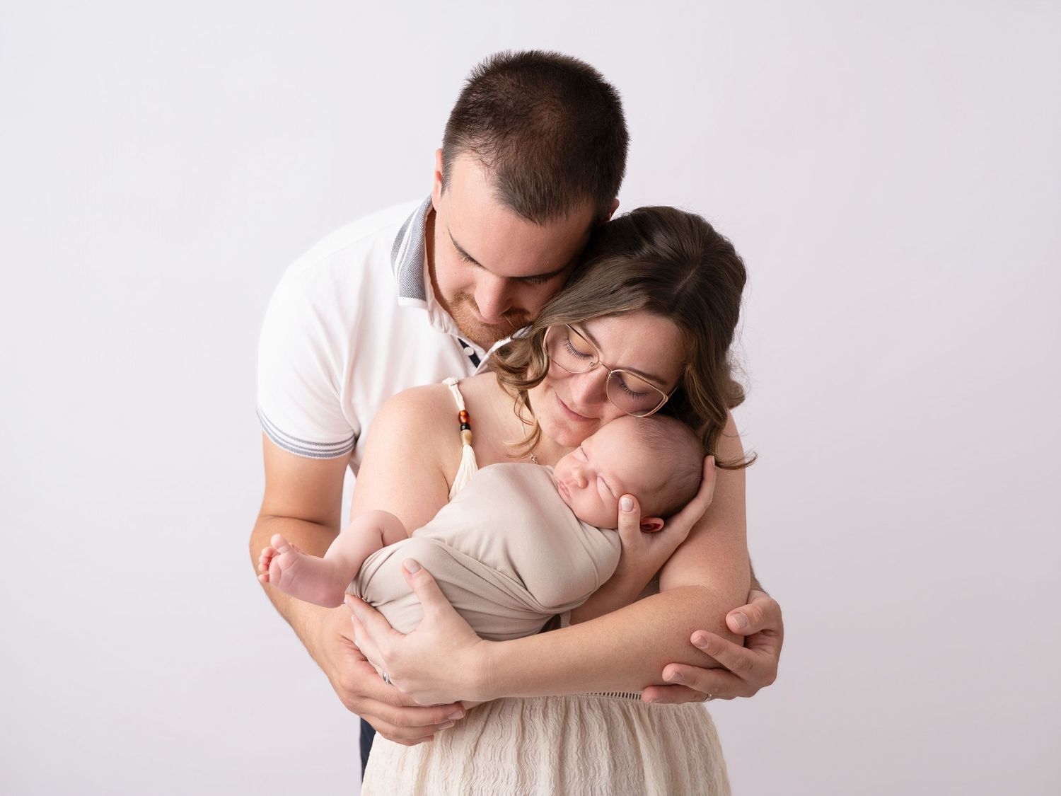 A couple tenderly embraces their sleeping newborn baby in a soft, neutral-toned professional studio portrait.