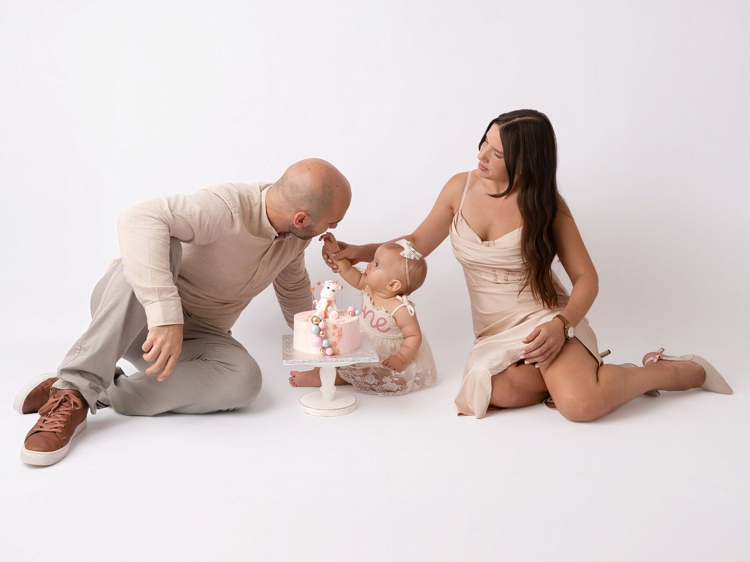 Family of three sits on white floor as parents watch their baby girl enjoy a small pink first birthday smash cake.