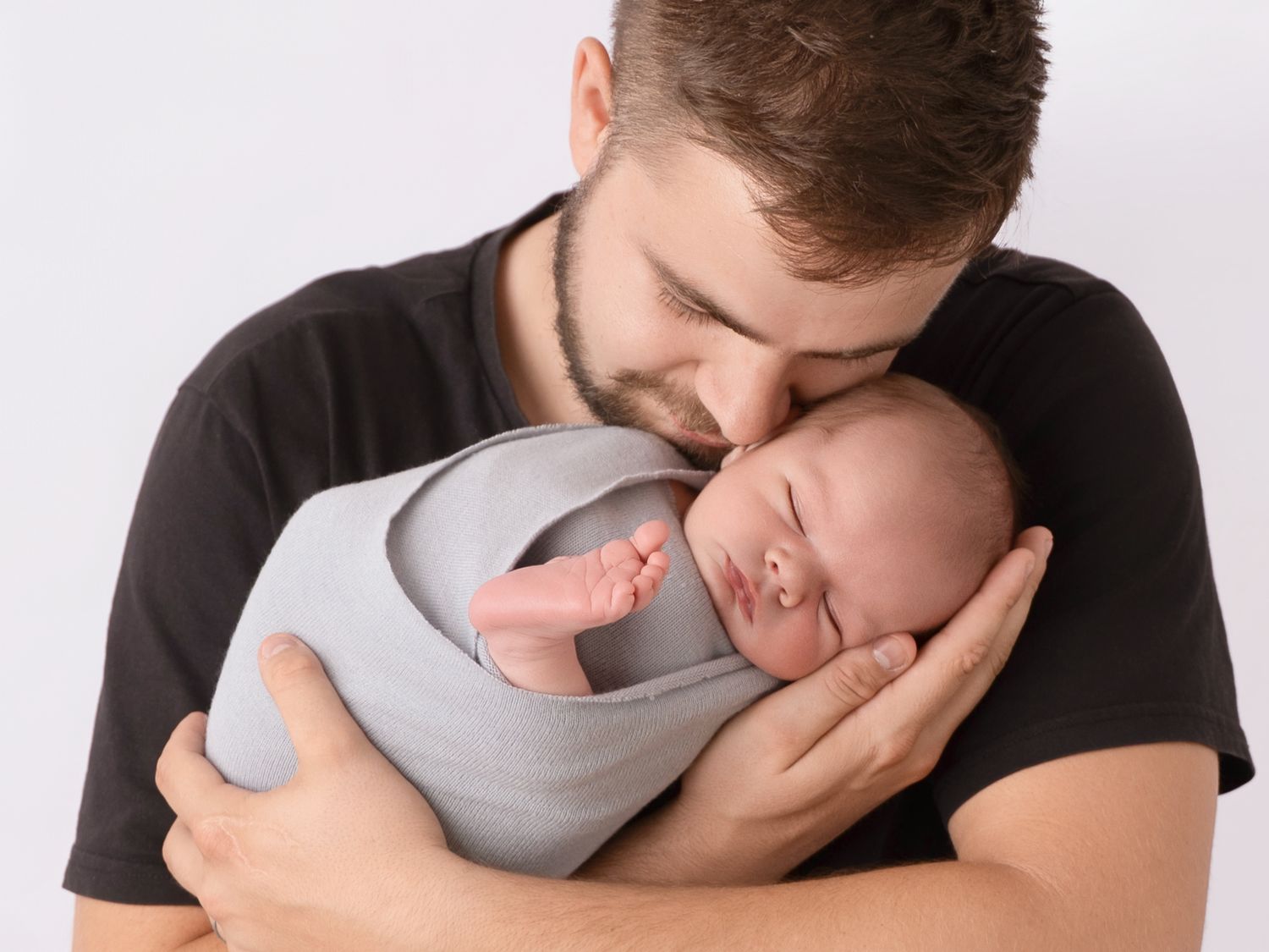 A loving father gently kisses his sleeping newborn baby wrapped in a soft gray swaddle against a white background.