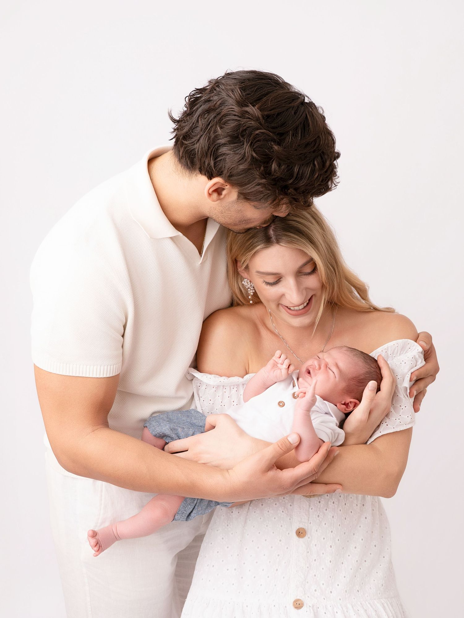 A couple in white outfits tenderly holds their newborn baby, with the father kissing the mother's head in a studio.