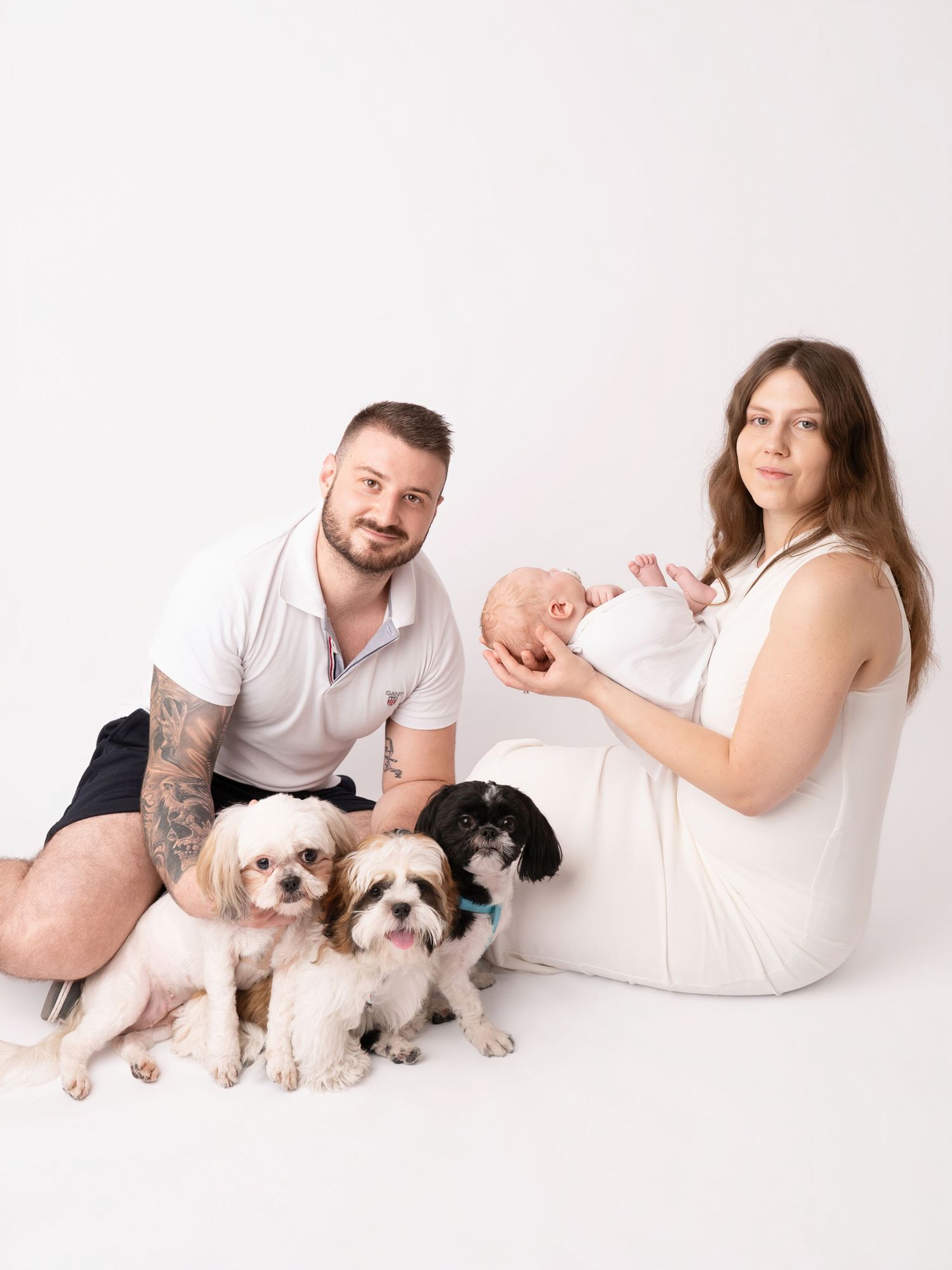 Family portrait with parents, newborn baby, and three small dogs posing together on a white studio background.