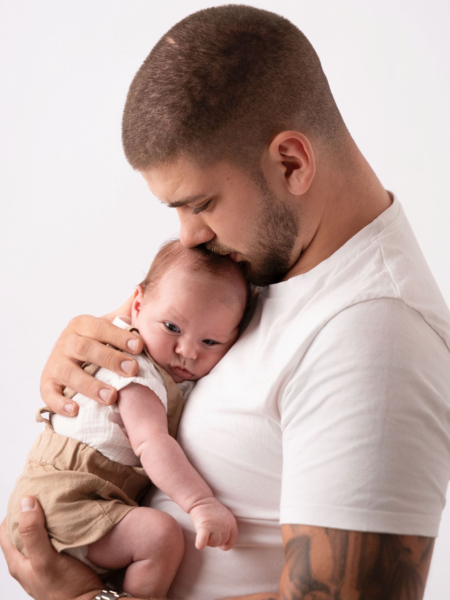 A tattooed father tenderly kisses his newborn baby while cradling the infant against his chest in a white t-shirt.