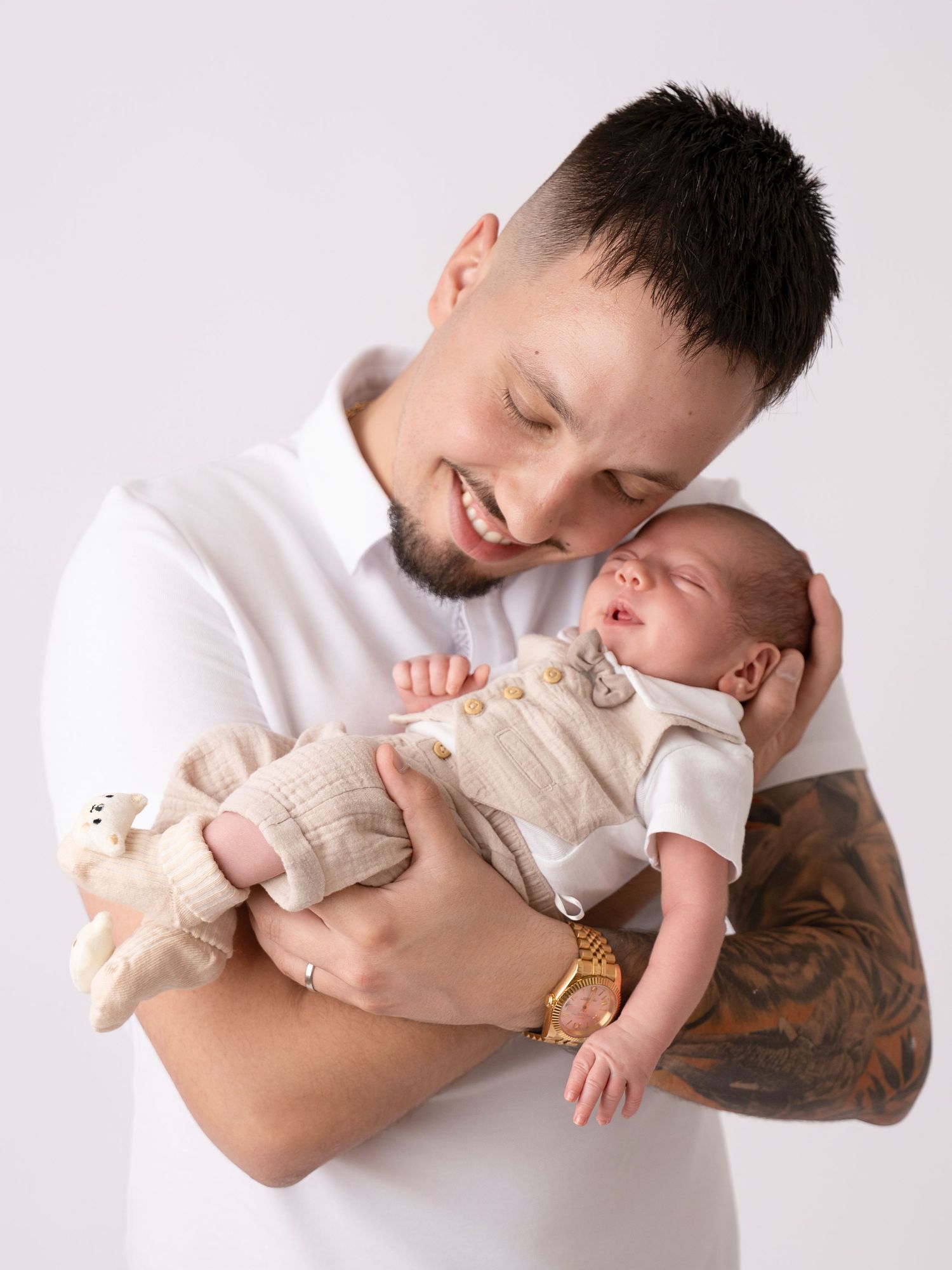 Smiling father with tattoos holding his sleeping newborn baby in a white studio setting.
