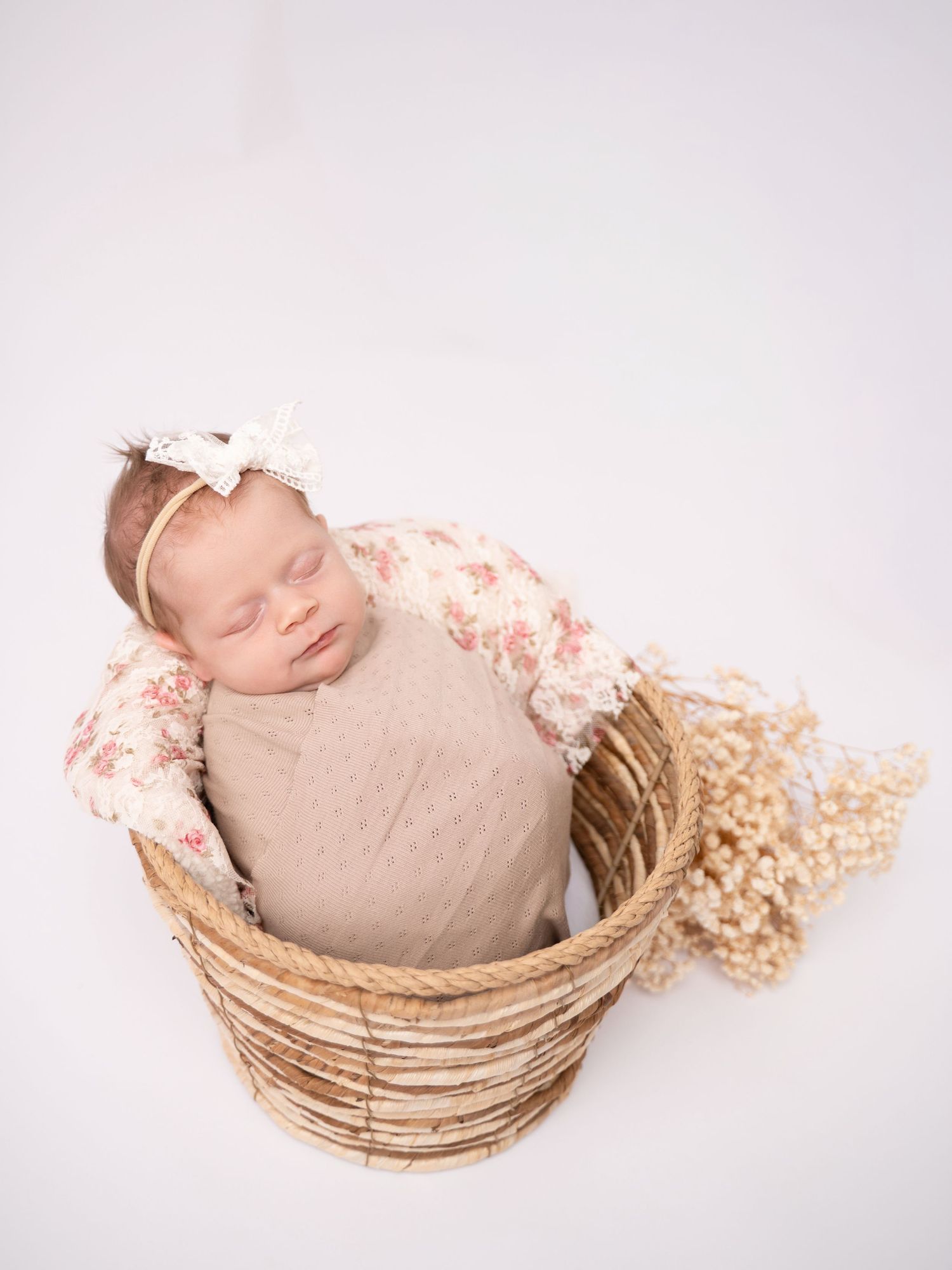 A sleeping newborn wrapped in a floral swaddle sits in a wicker basket with dried flowers.