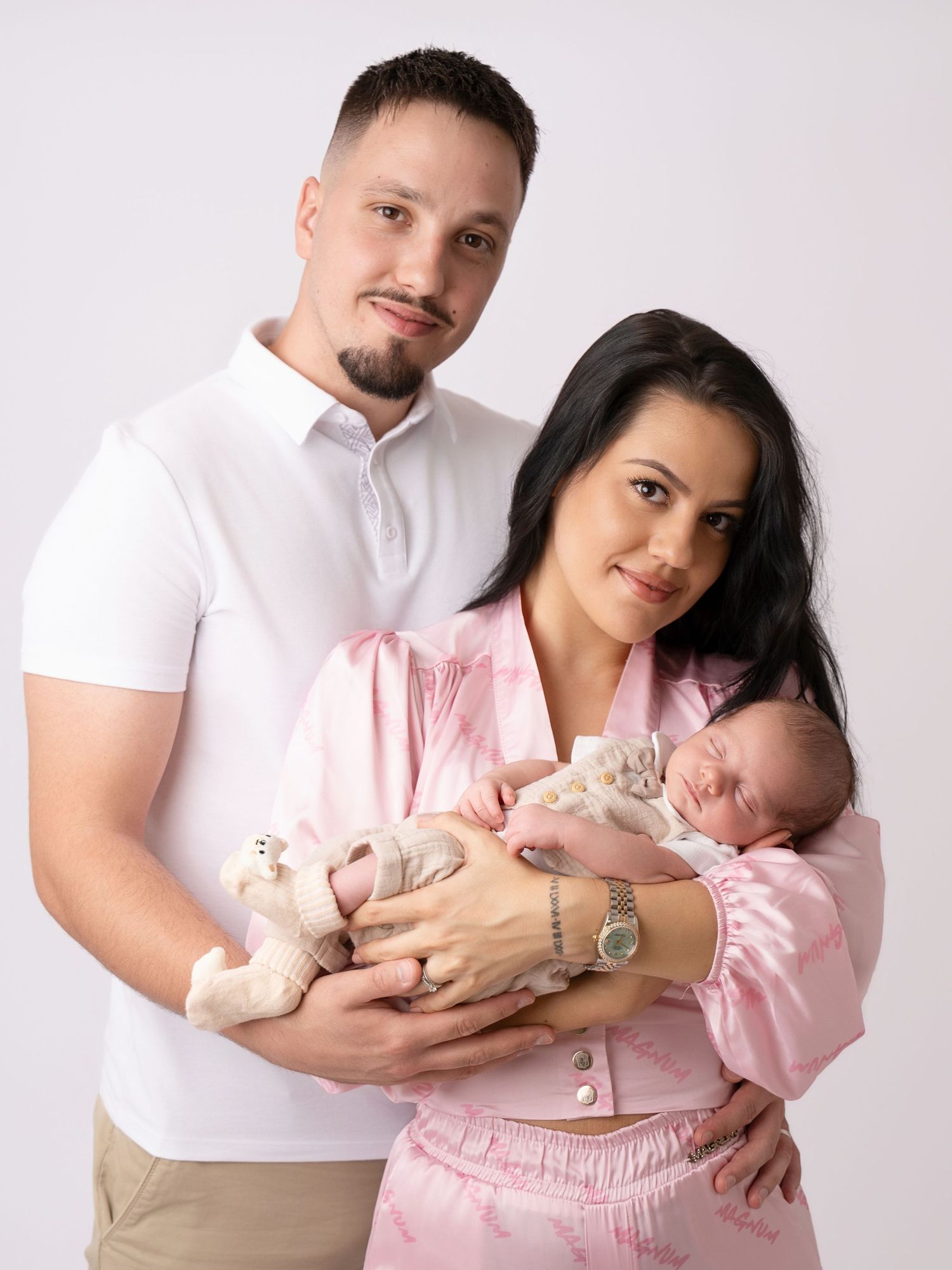 A couple in a studio portrait holding a newborn baby and a small teddy bear, wearing white and pink outfits.