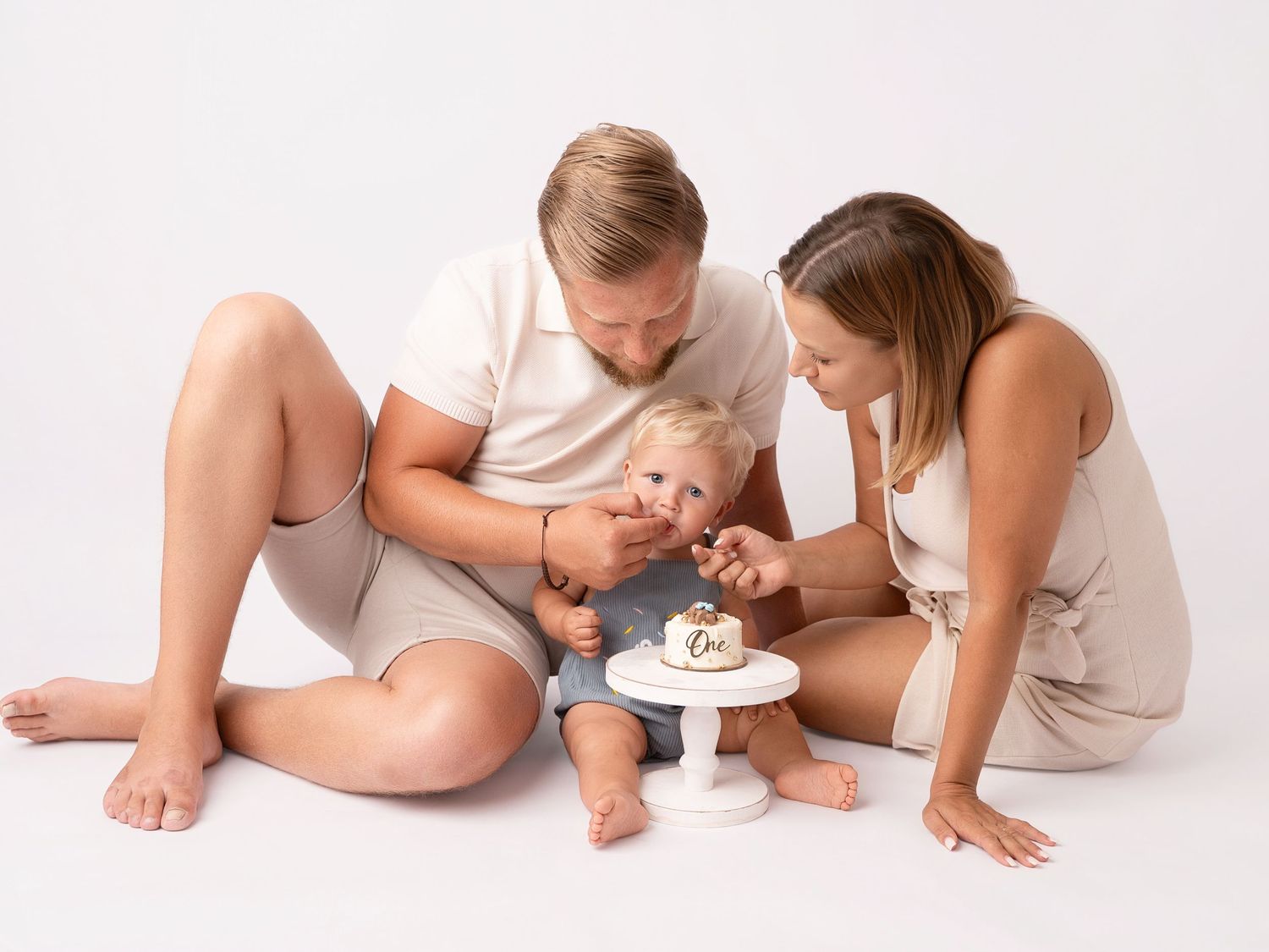 Family celebrating baby's first birthday with a smash cake on a white background.