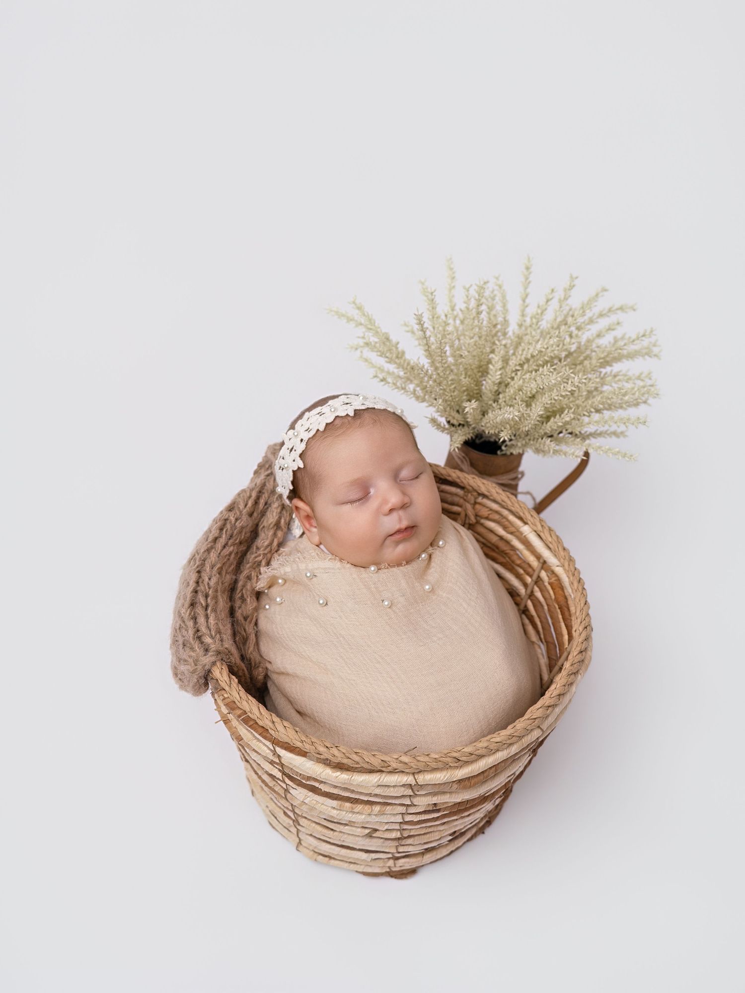 Sleeping newborn baby wrapped in beige swaddle with lace headband, nestled in wicker basket with dried greenery.