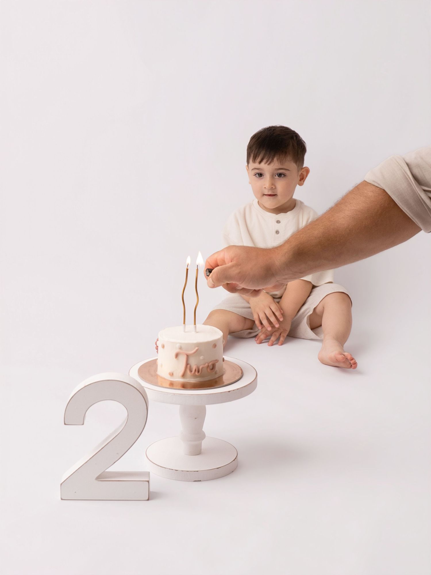 Toddler boy sitting beside a birthday cake on white stand with number 2, as adult lights candles.