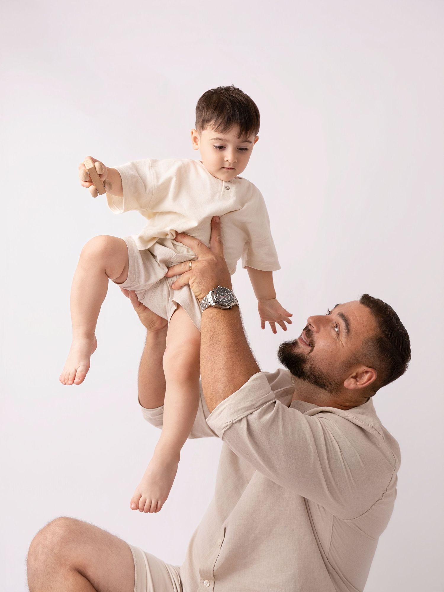 A bearded man in beige hoodie lifts a smiling toddler in cream outfit against white background.