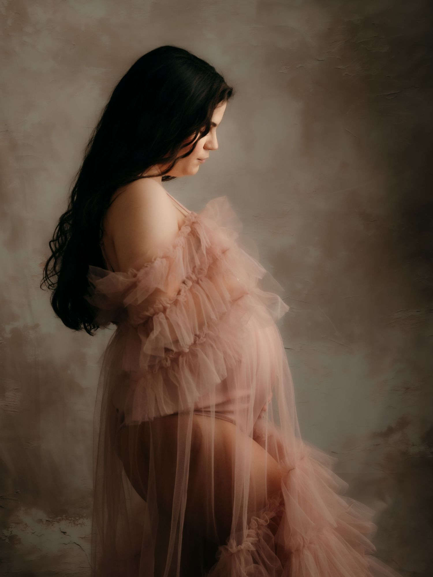 Pregnant woman in a sheer blush ruffled tulle maternity gown looking down in a softly lit studio portrait.