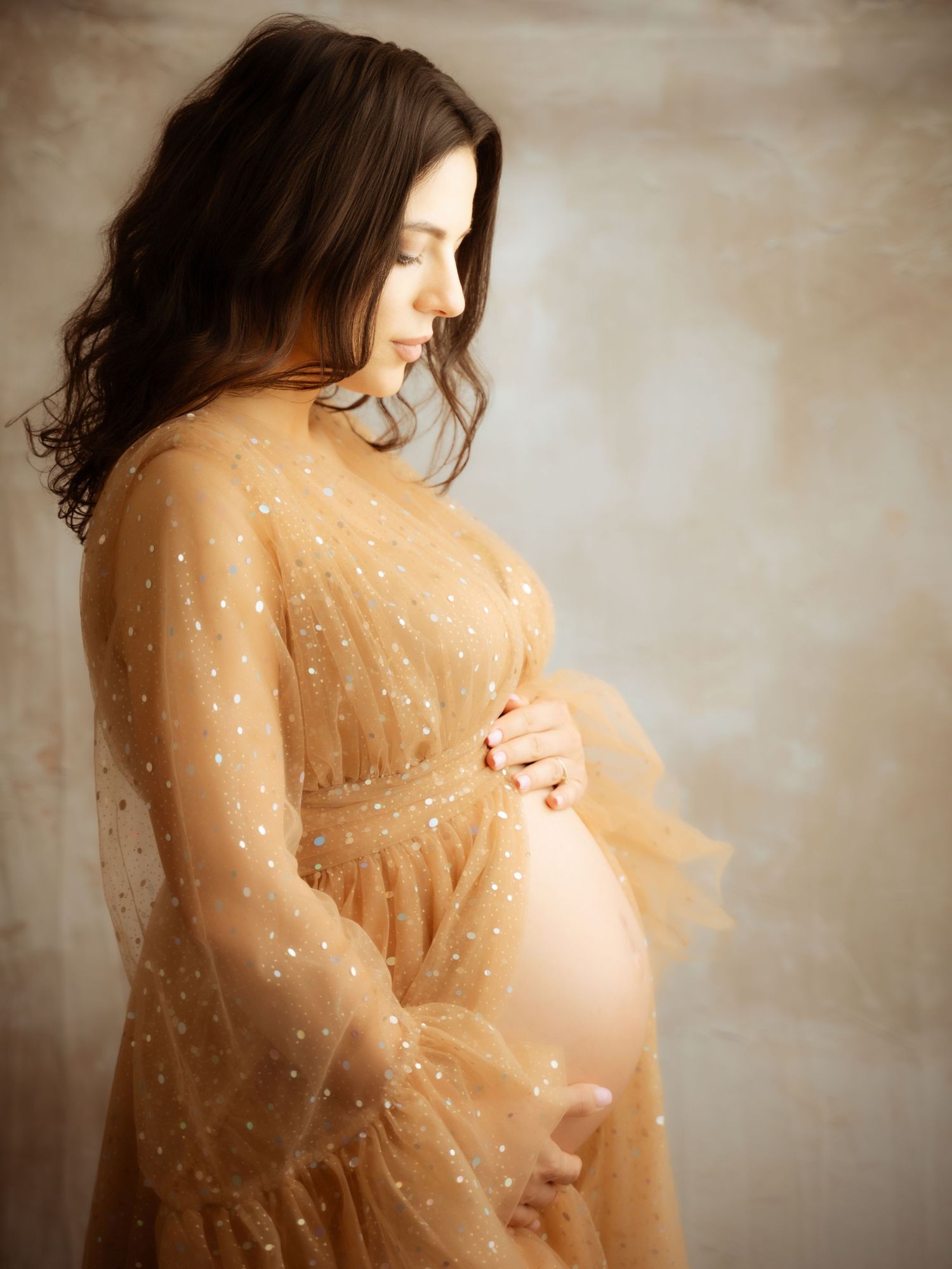 Side profile of pregnant woman in gold tulle dress cradling belly.