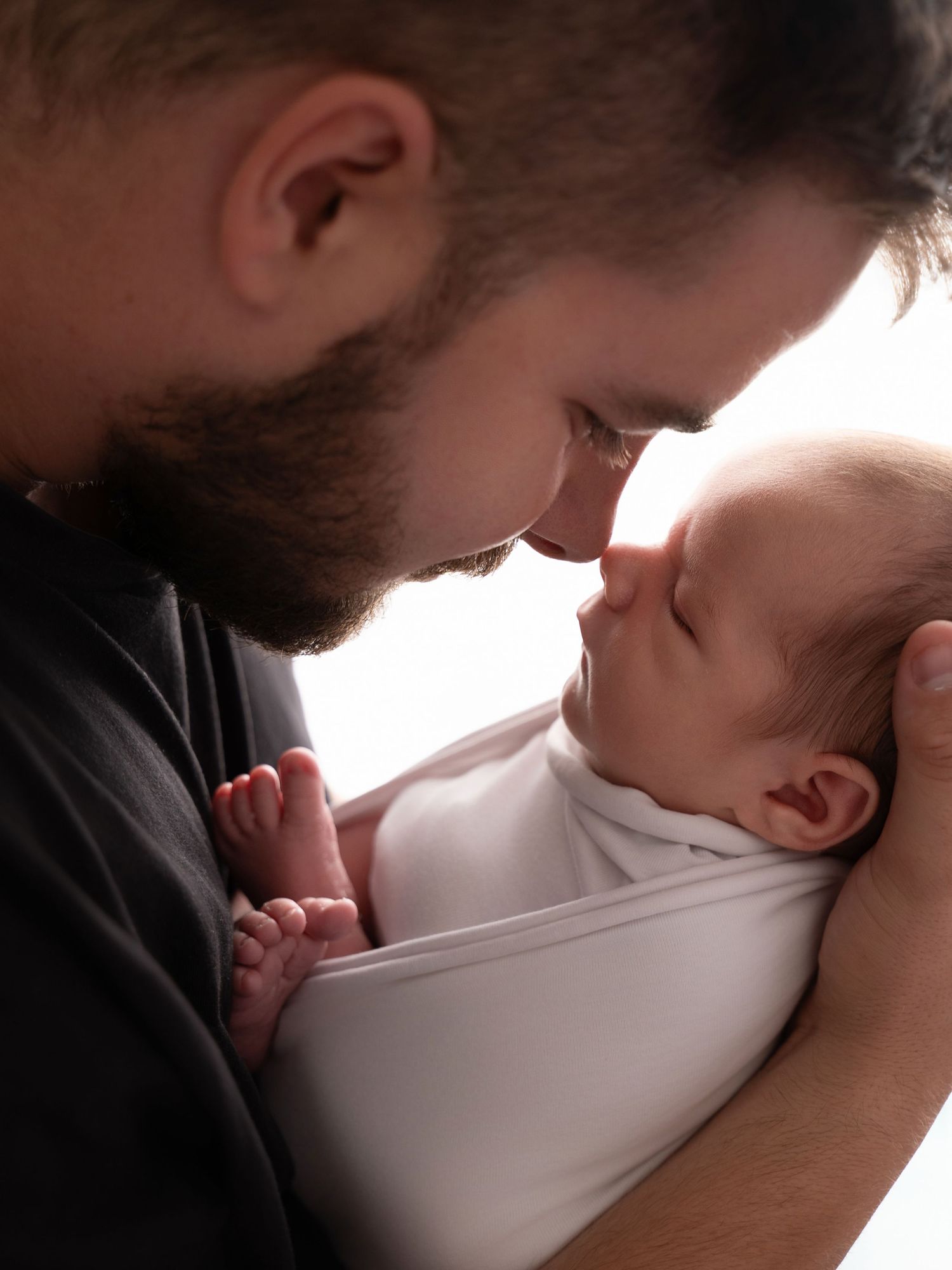 Bearded father tenderly touches noses with swaddled newborn baby while cradling infant in his arms.