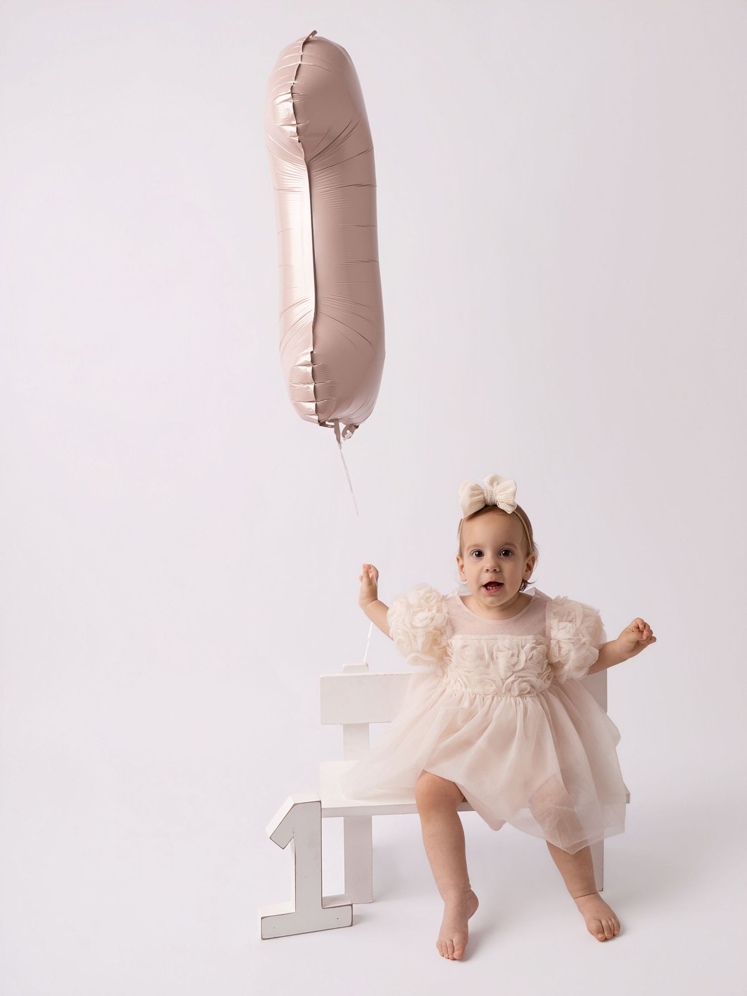 Baby girl in white tulle dress holds pink number 1 balloon, celebrating her first birthday milestone.