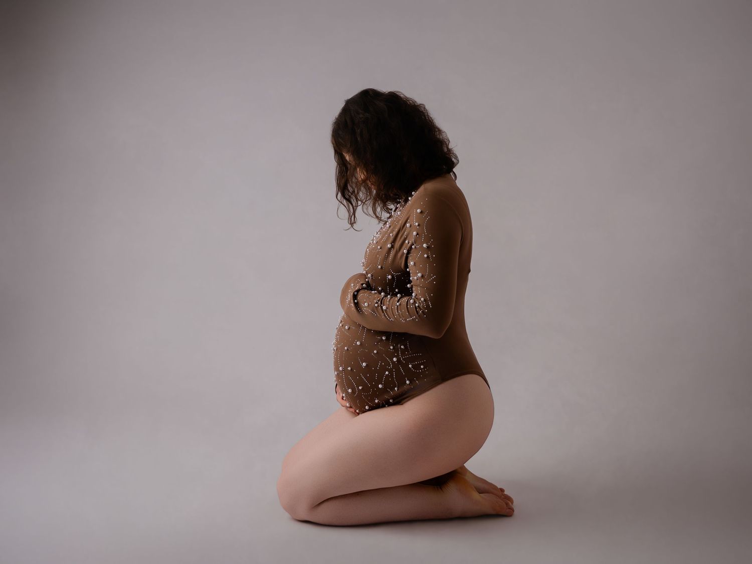 Pregnant woman in a lace bodysuit kneeling on the floor, cradling her baby bump in a serene maternity photo.