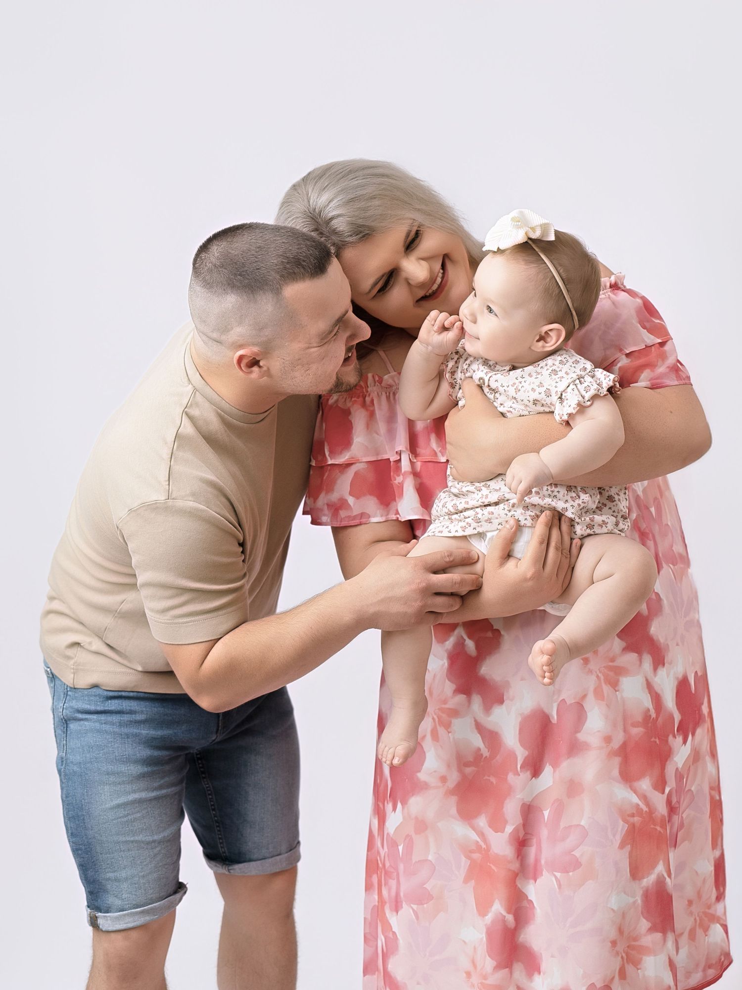 Loving parents lean in to kiss their baby girl in a floral dress during a studio photoshoot.