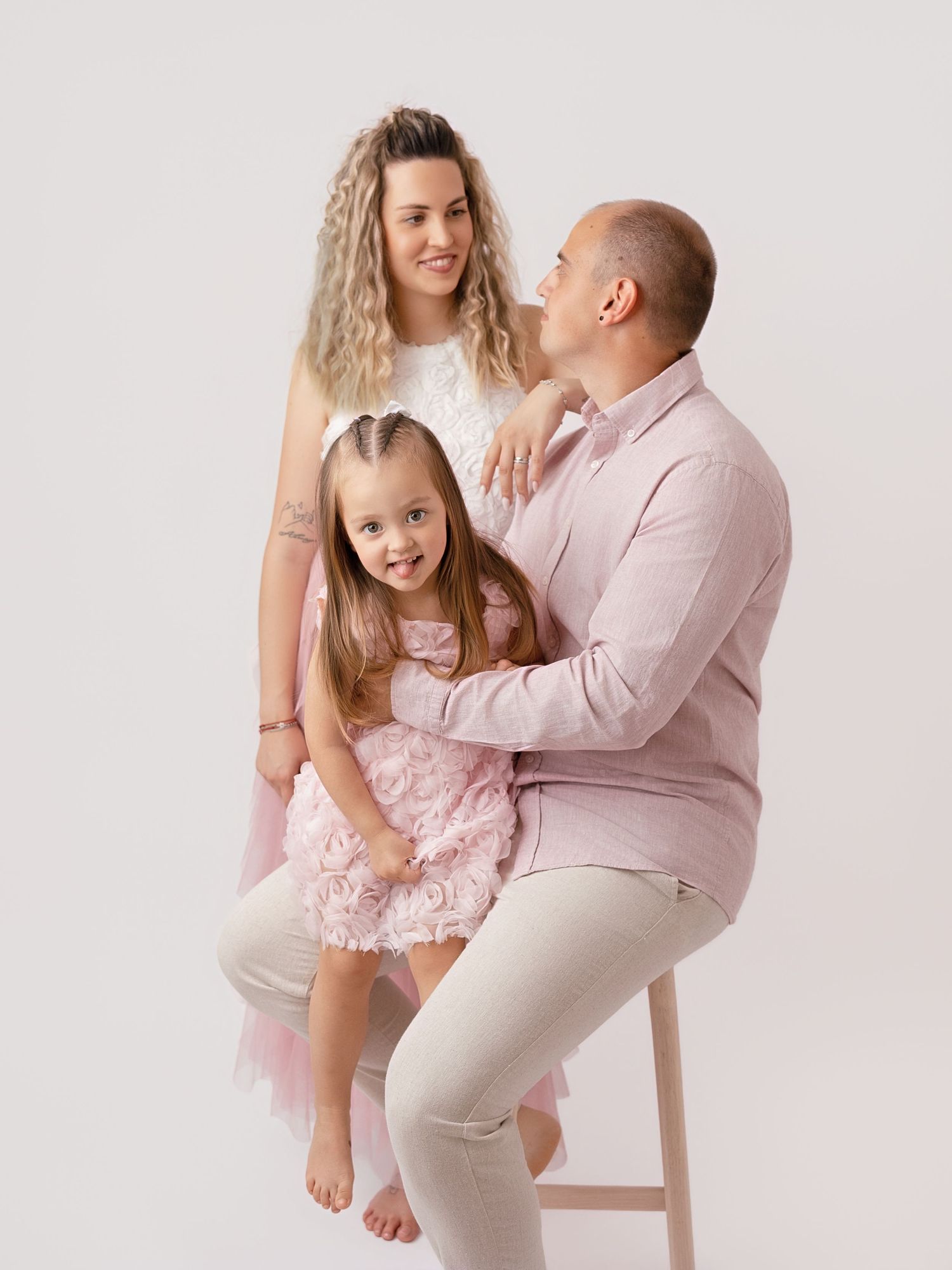 A family of three in pink outfits poses for a studio portrait, with parents and young daughter smiling warmly.