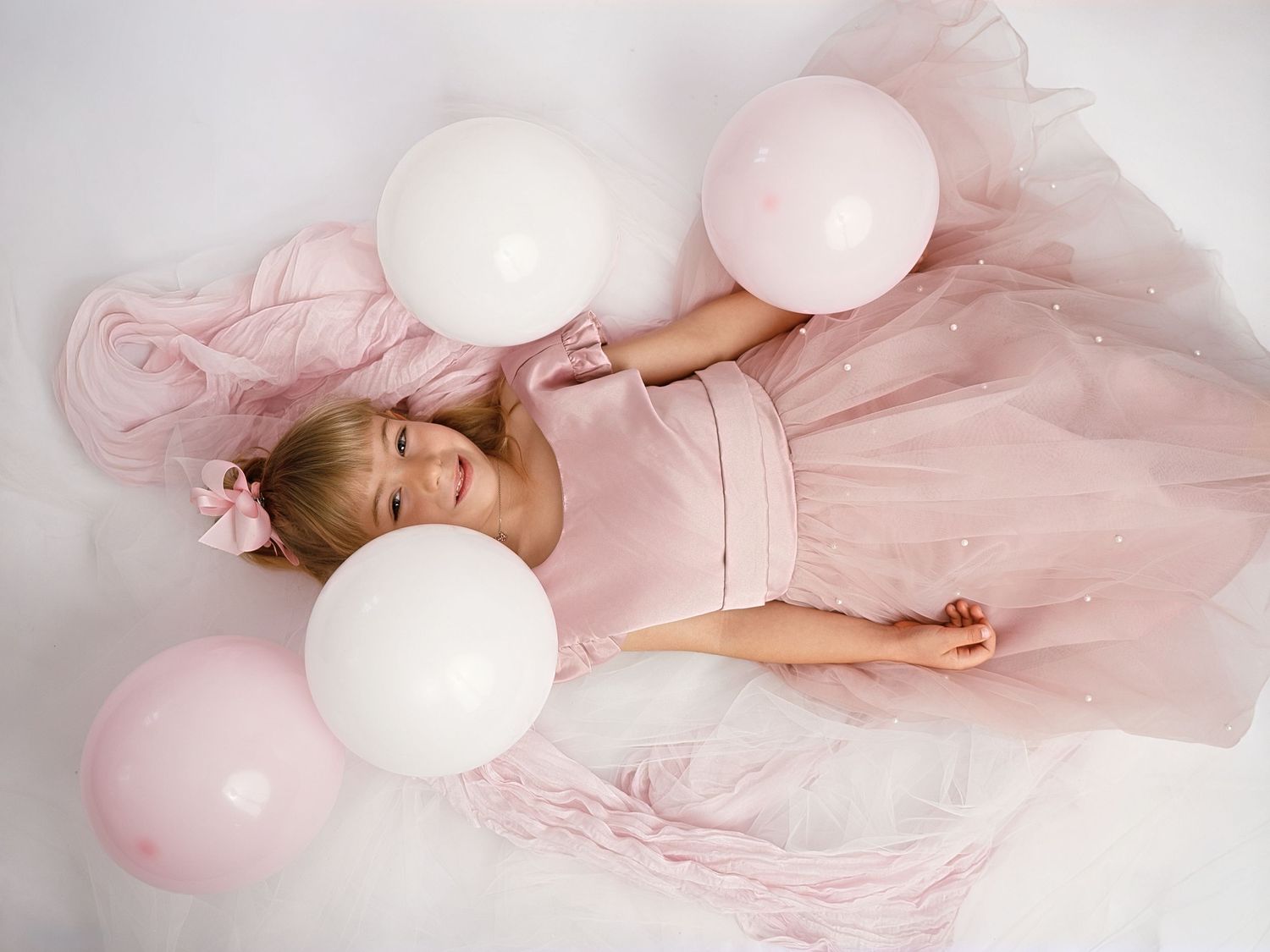 Young girl in pink tulle dress lying among white and pink balloons on white background.
