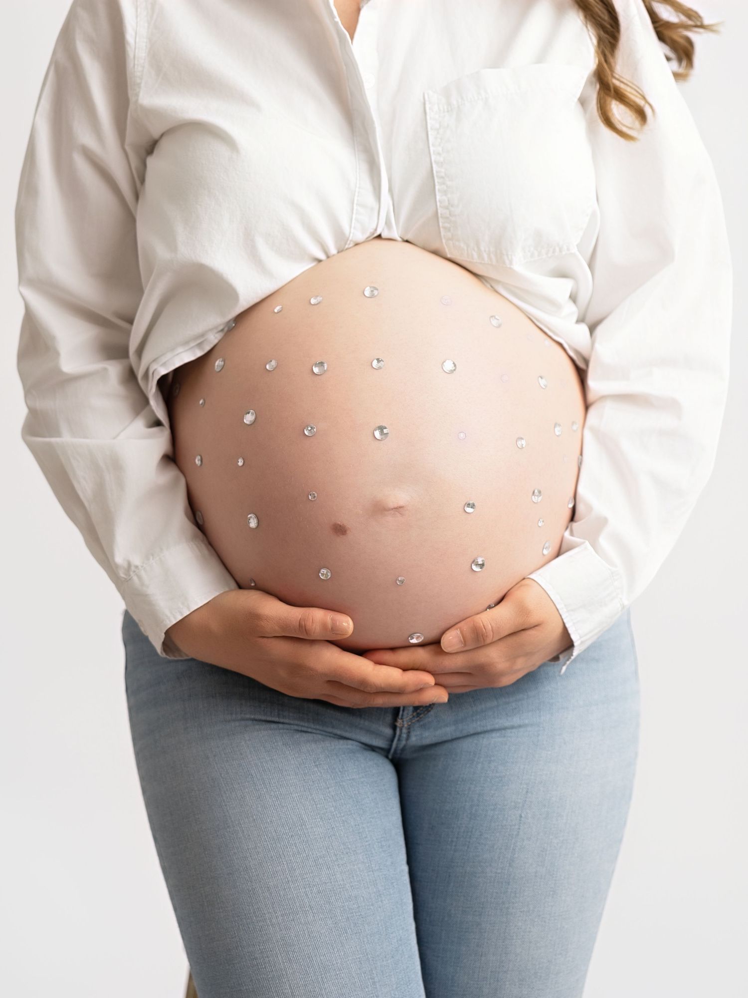 Close-up of pregnant woman's bare baby bump adorned with sparkling rhinestone gems, wearing white shirt and jeans.