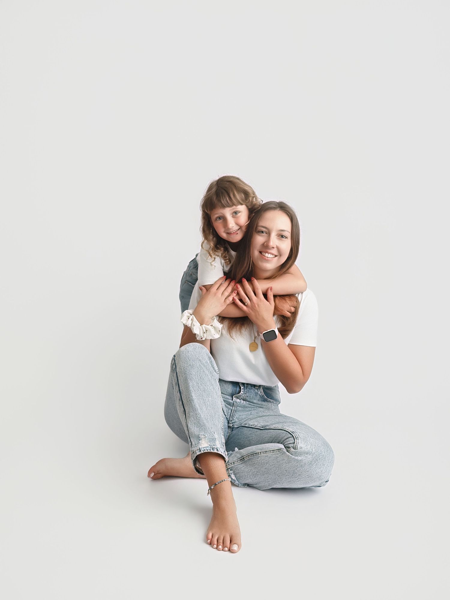 Two girls in jeans hugging and smiling on a white background, one sitting cross-legged while the other embraces from behind.