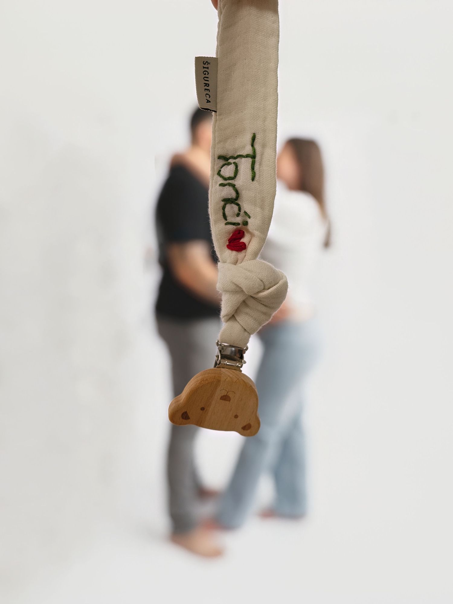 A personalized baby pacifier clip with the name Toni embroidered on fabric, with a couple embracing in the blurred background.