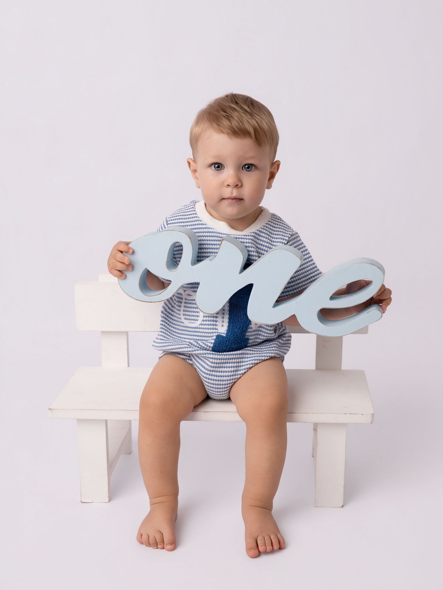 A baby boy sitting on a white bench holding a light blue cursive “one” sign, celebrating his first birthday.