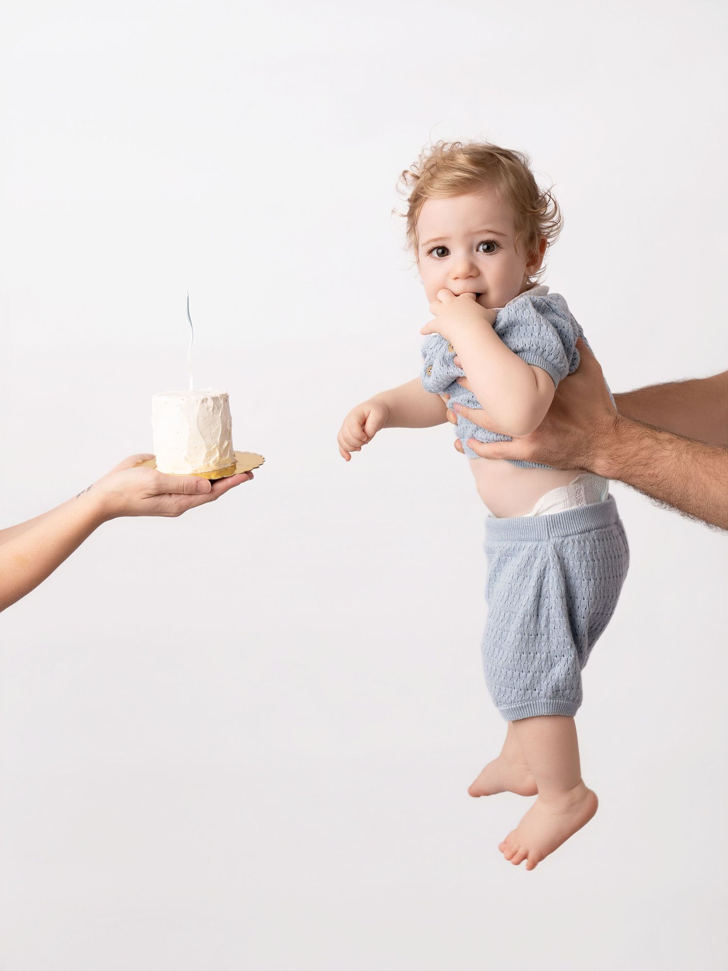 A curious baby in a blue knit romper reaches toward a small birthday cake held by an adult hand on a white background.