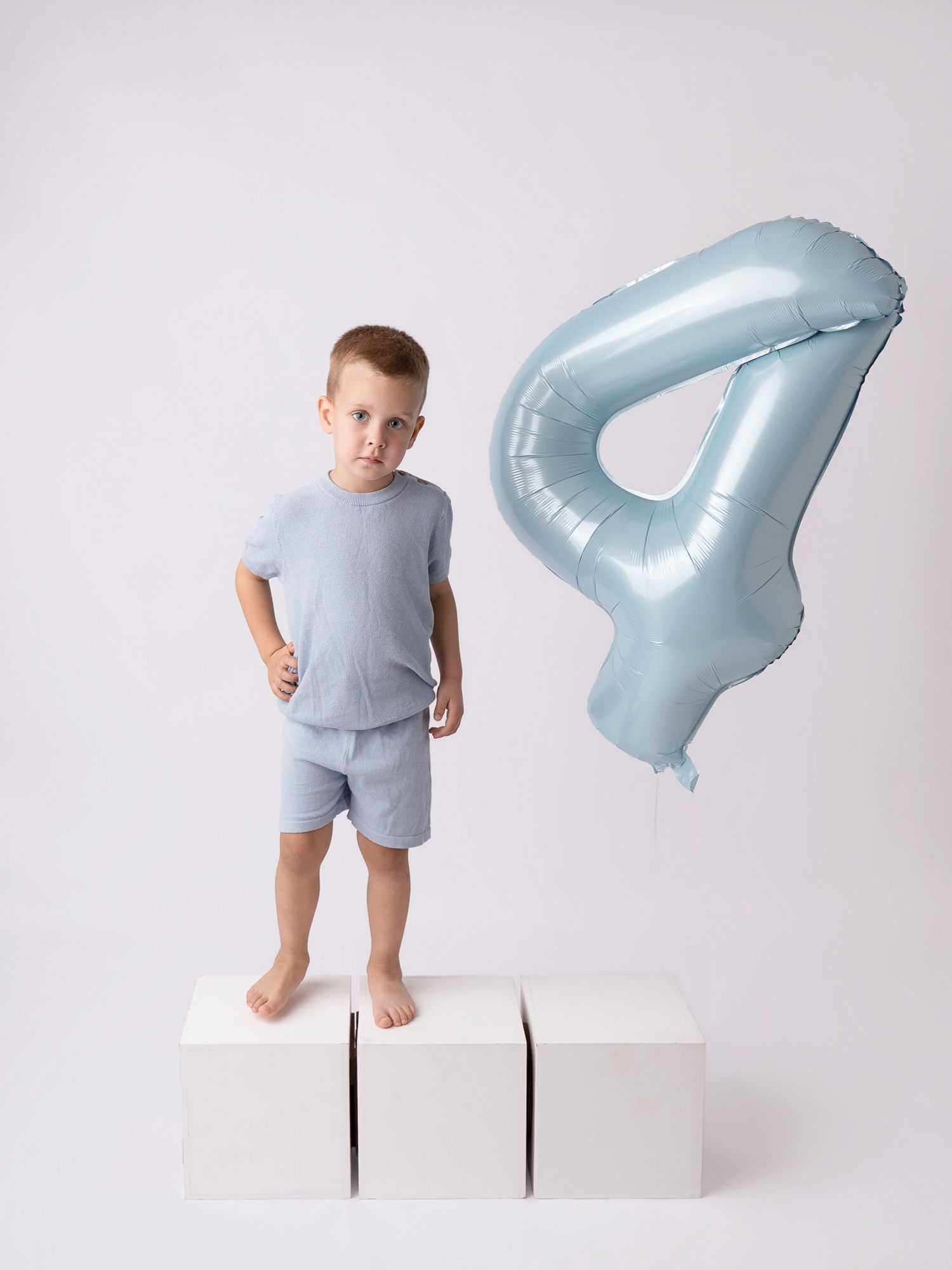 A young boy in light blue outfit stands on white blocks beside a number 4 balloon, celebrating his 4th birthday.