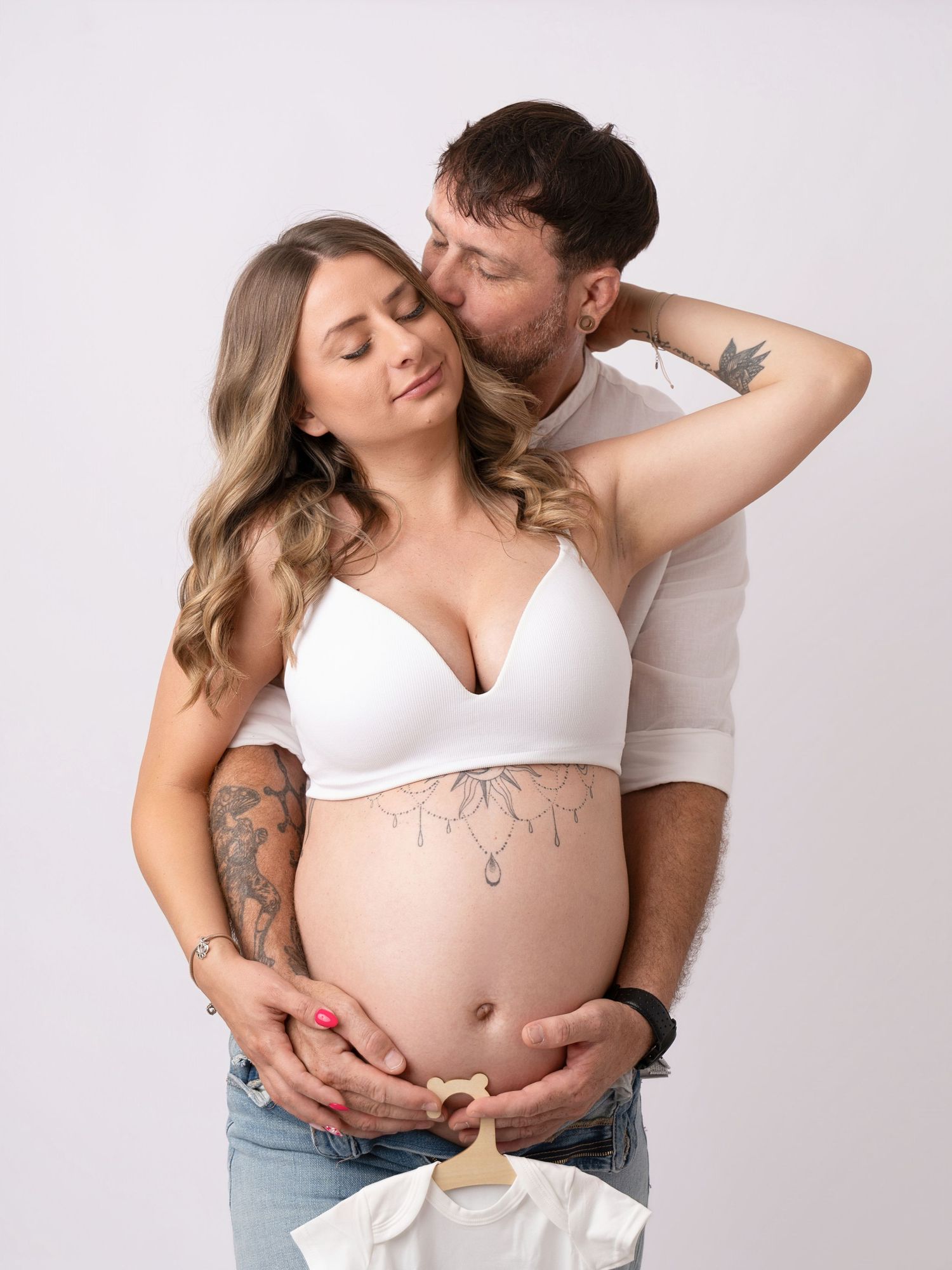 Couple embracing during maternity shoot, man kissing pregnant woman holding baby clothes.