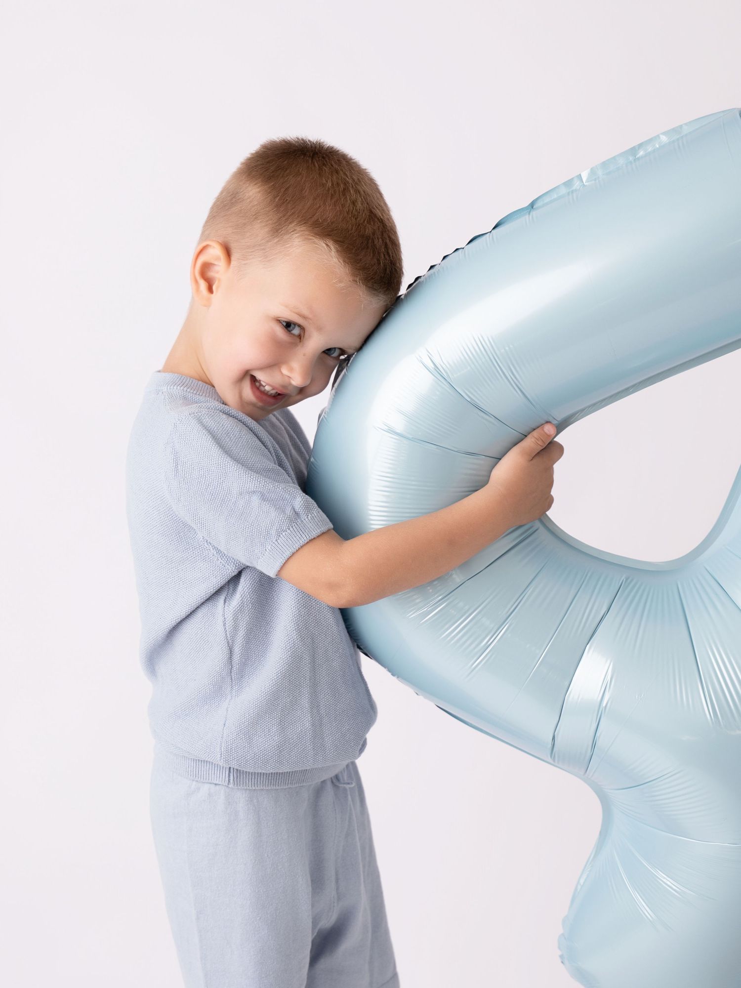 Young boy in gray outfit hugging a large light blue number balloon, smiling against white background.