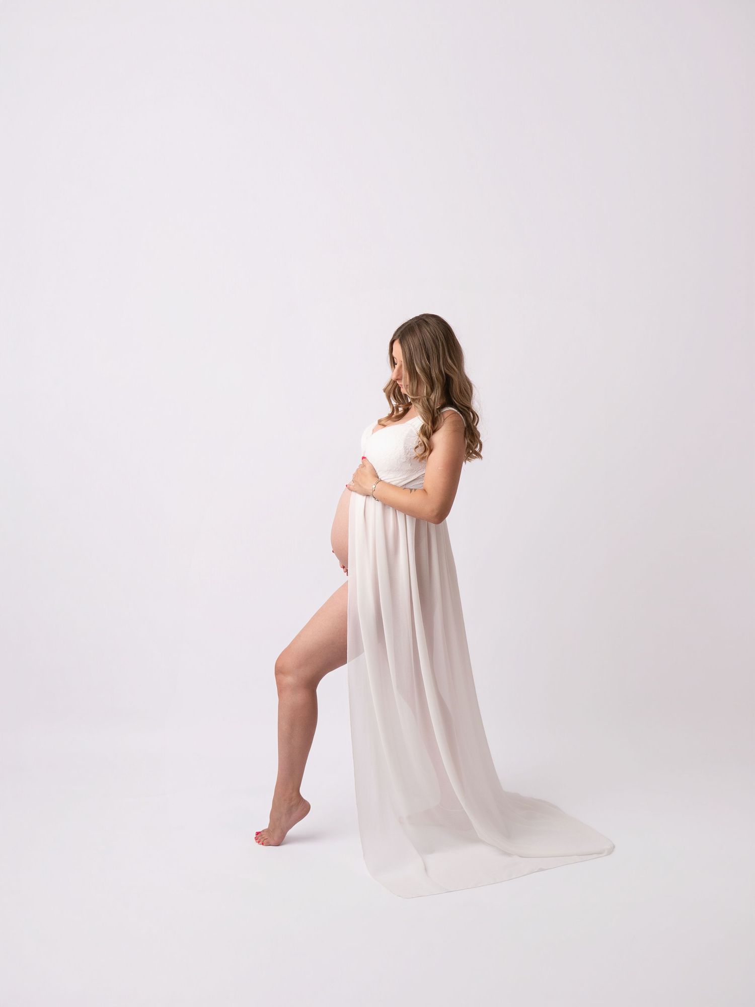 Pregnant woman in white flowing maternity gown posing sideways, cradling baby bump in a studio setting.