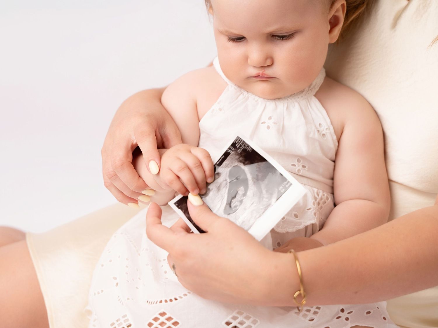 Baby girl in white dress holding an ultrasound photo while sitting on mother's lap during maternity photoshoot.