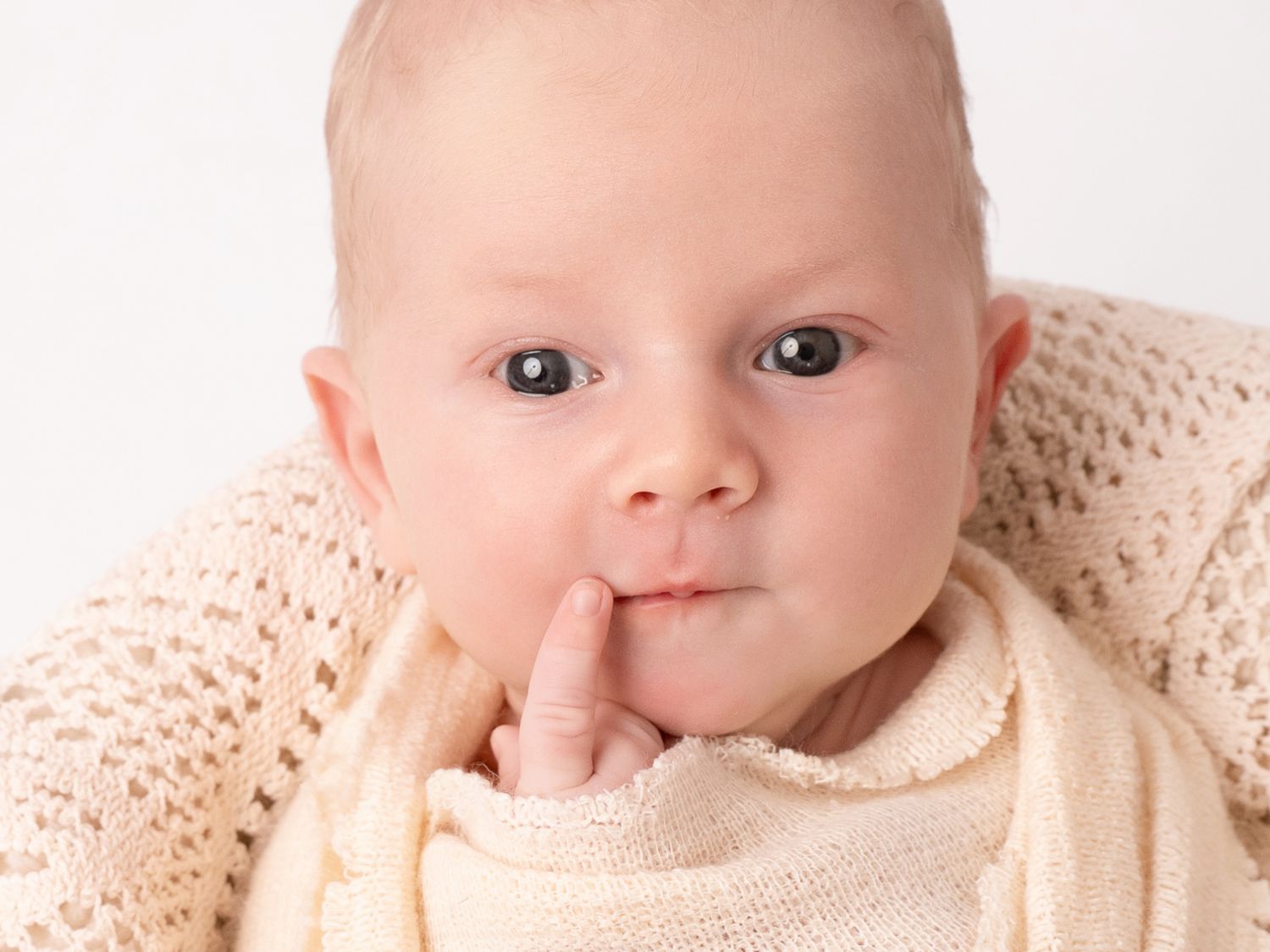Adorable baby in cream knit sweater with finger on lips against white background.