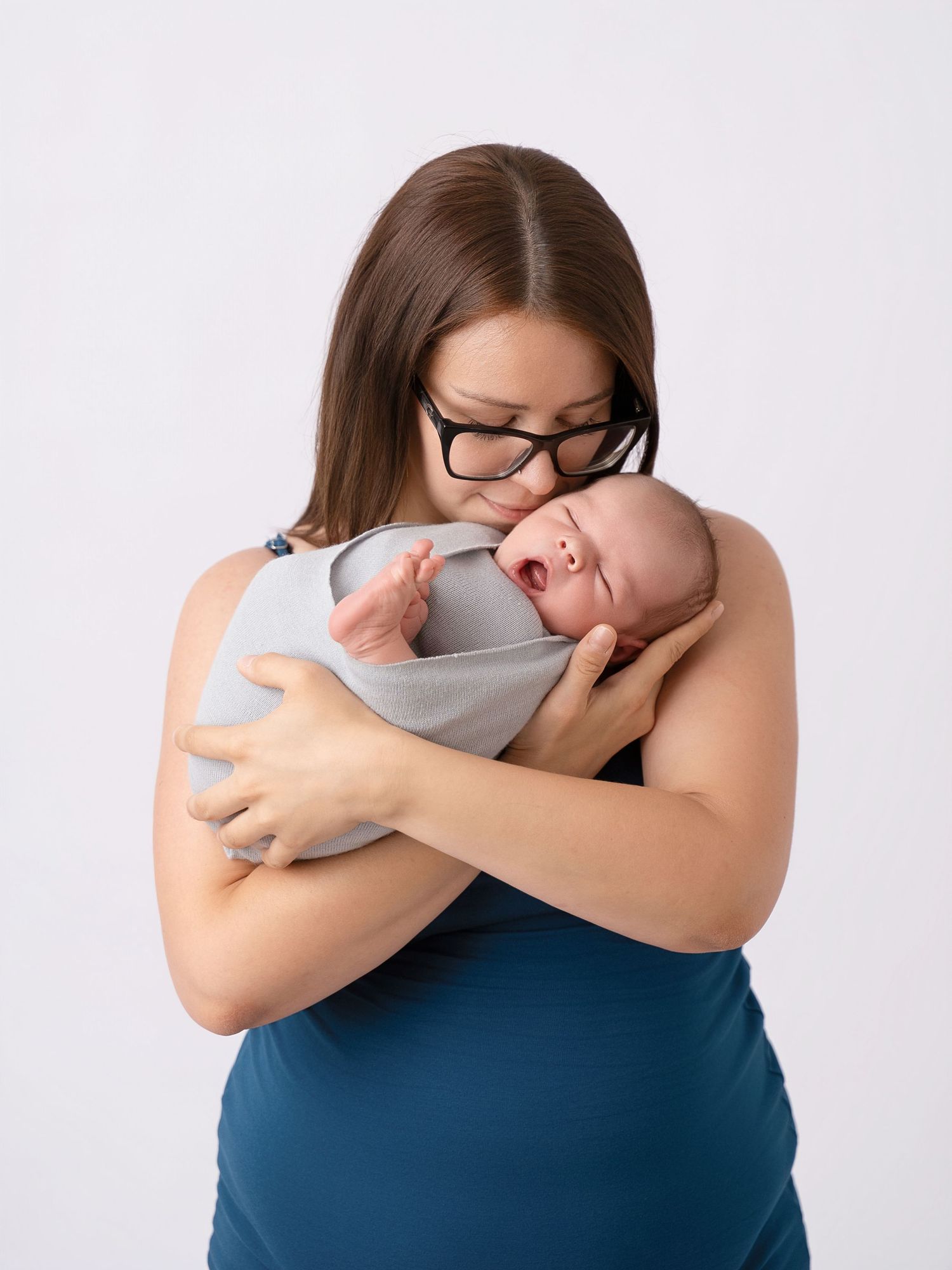 Dark-haired mother wearing glasses gently kisses her sleeping swaddled newborn baby on the head.