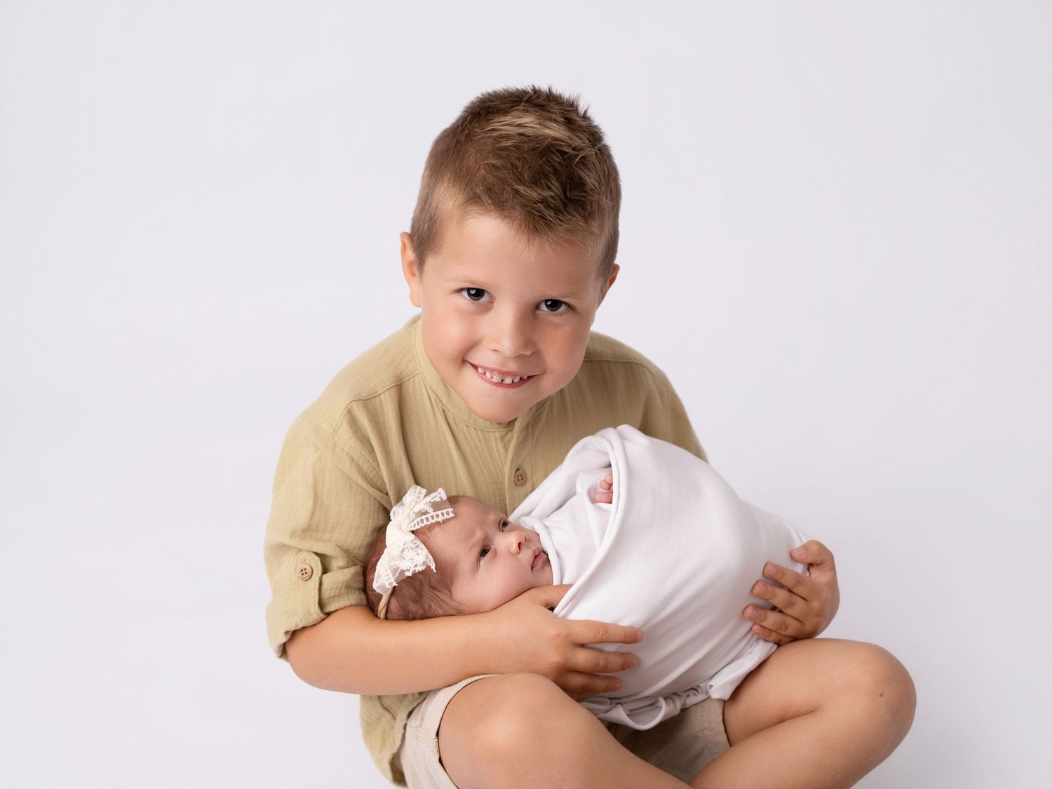 Smiling young boy sitting cross-legged holding swaddled newborn baby sibling on white background.