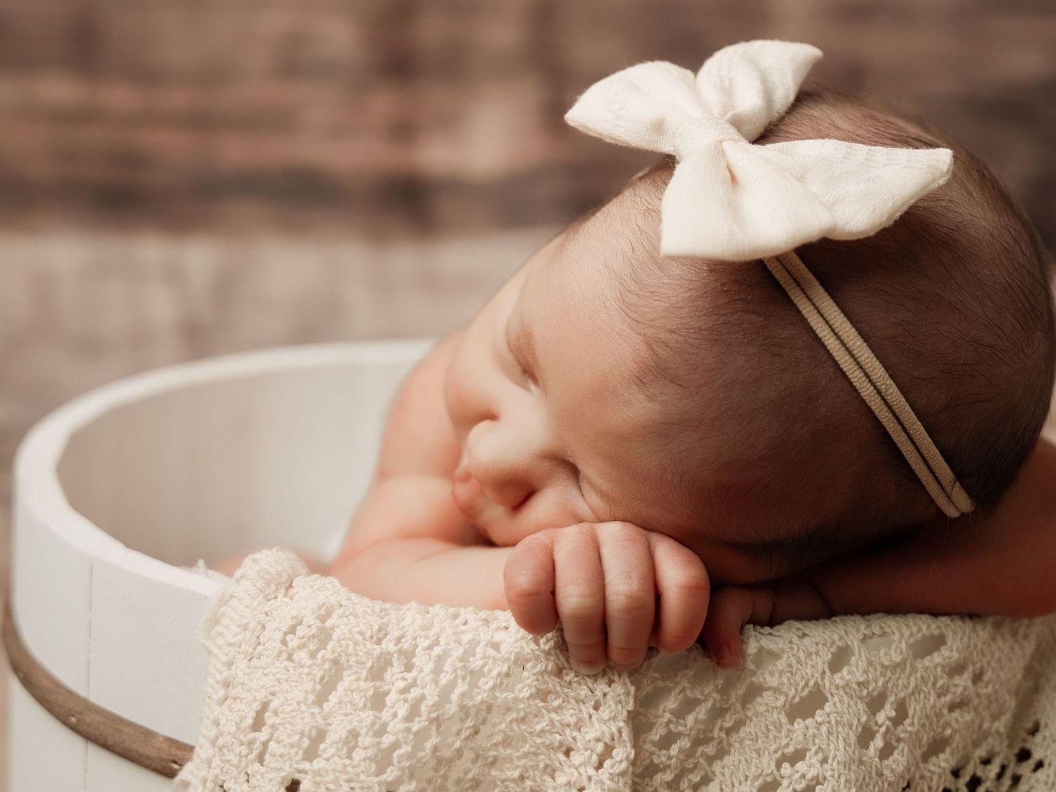 Peaceful newborn baby sleeping in a white wooden bucket, wearing a cream bow headband, wrapped in lace fabric.