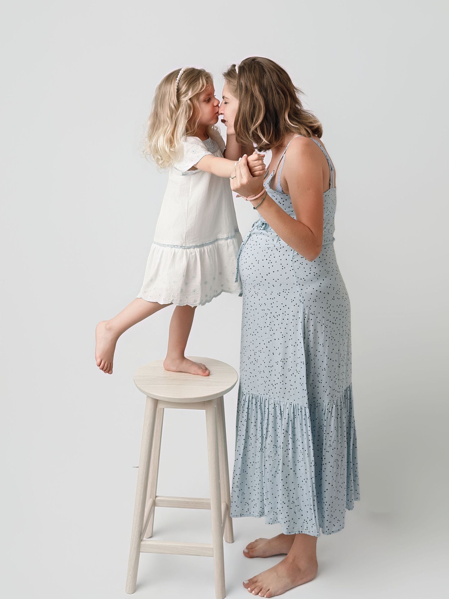 A mother and young daughter share a tender moment, dressed in soft floral and white dresses, standing by a wooden stool.