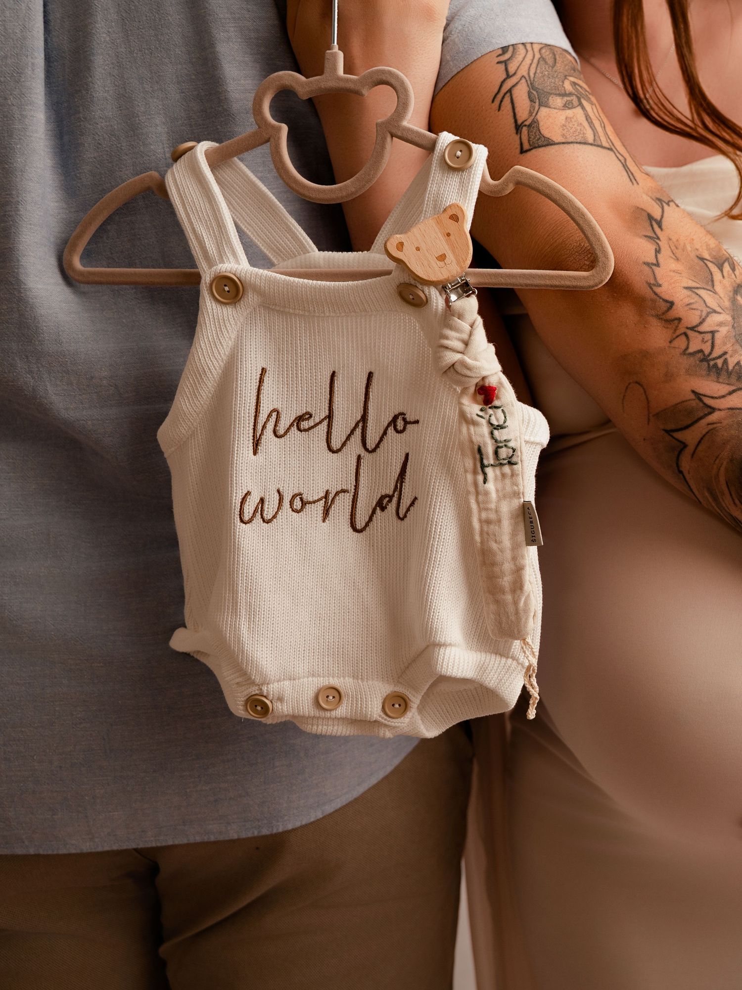 A white hello world baby romper on a bear hanger held by a tattooed couple during a maternity photoshoot.