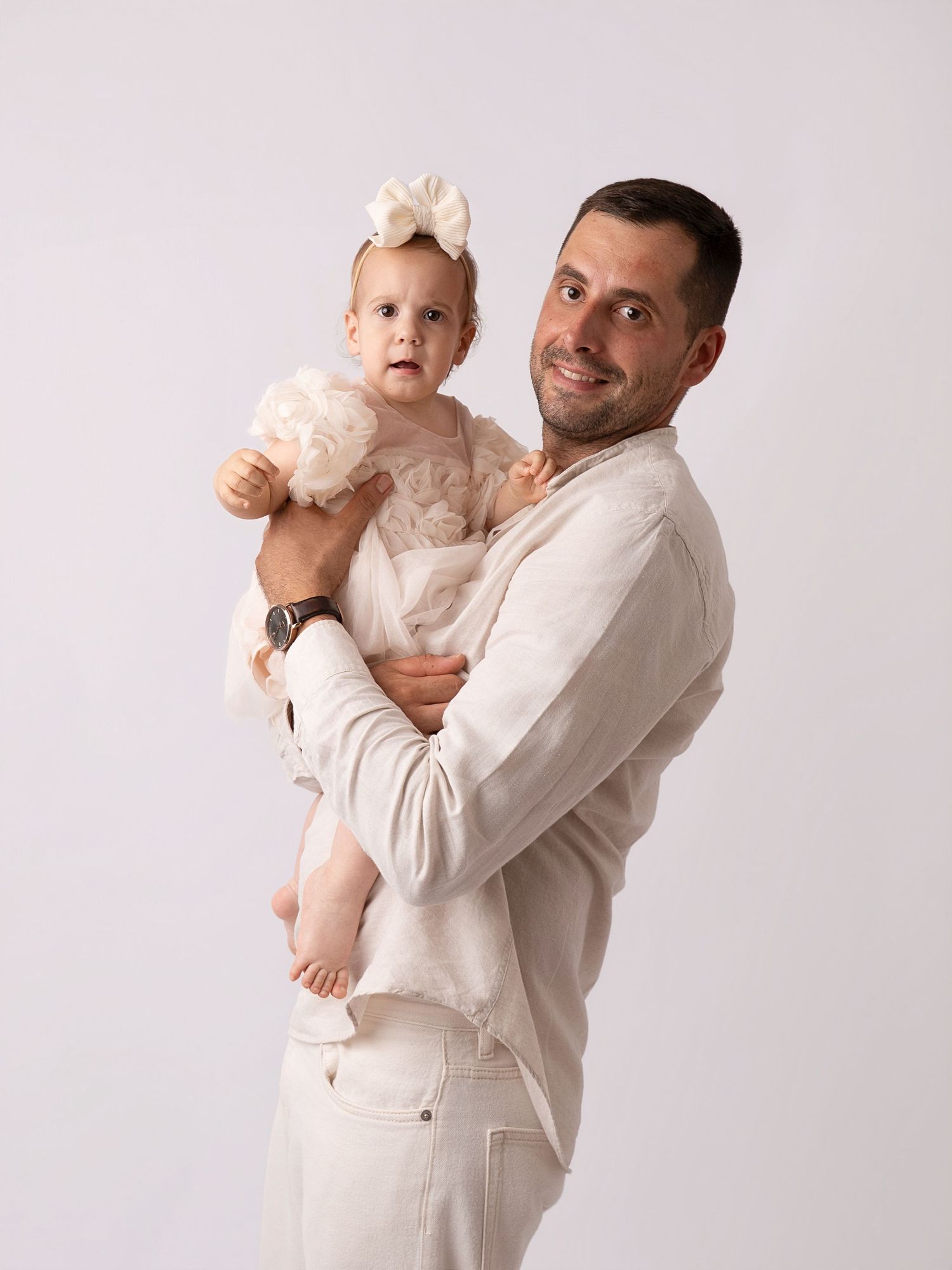 Smiling man in white outfit holding a baby girl in a white tulle dress with a bow headband.