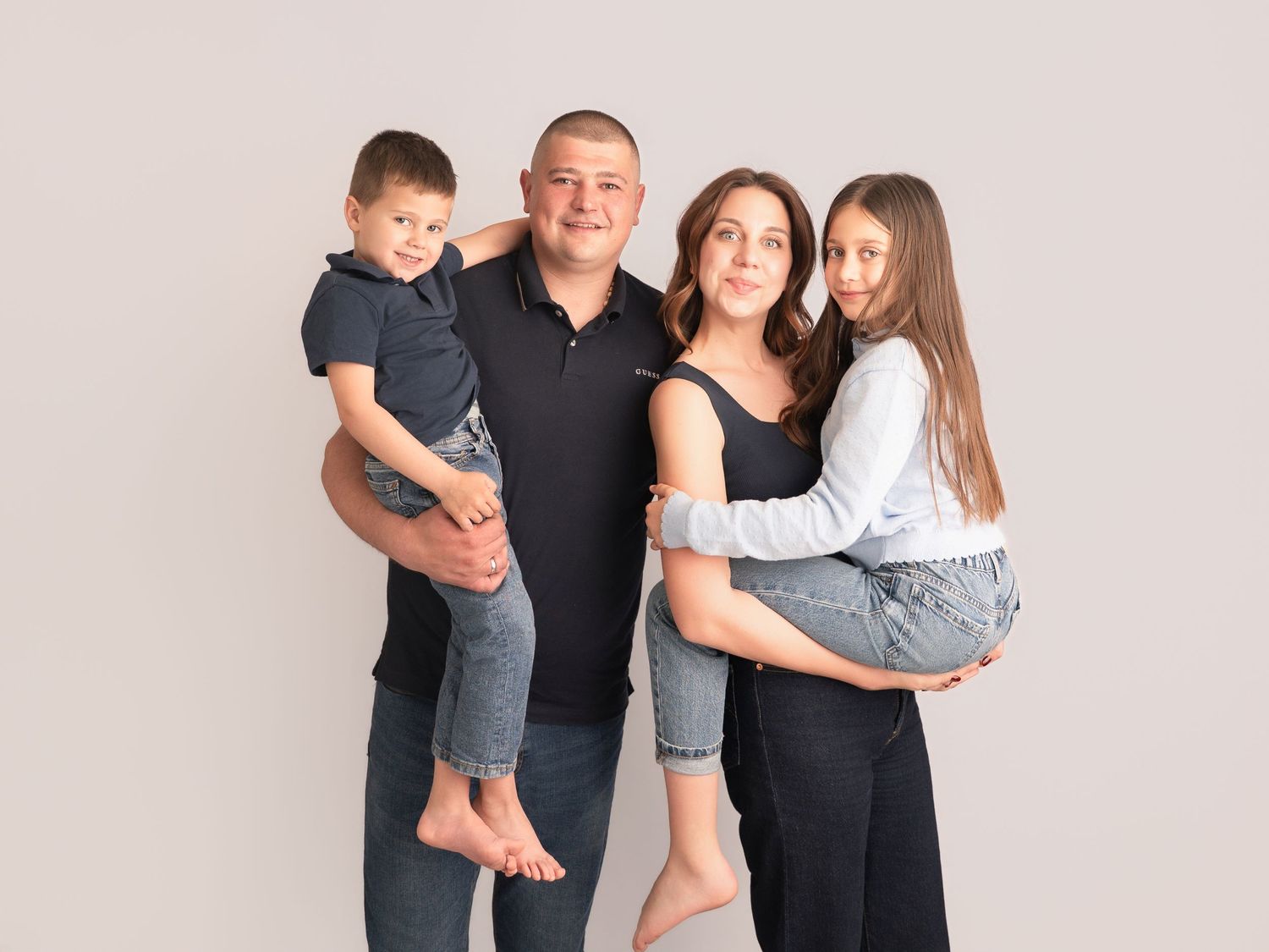 A happy family of four posing together, parents holding their two young children, against a light gray studio background.