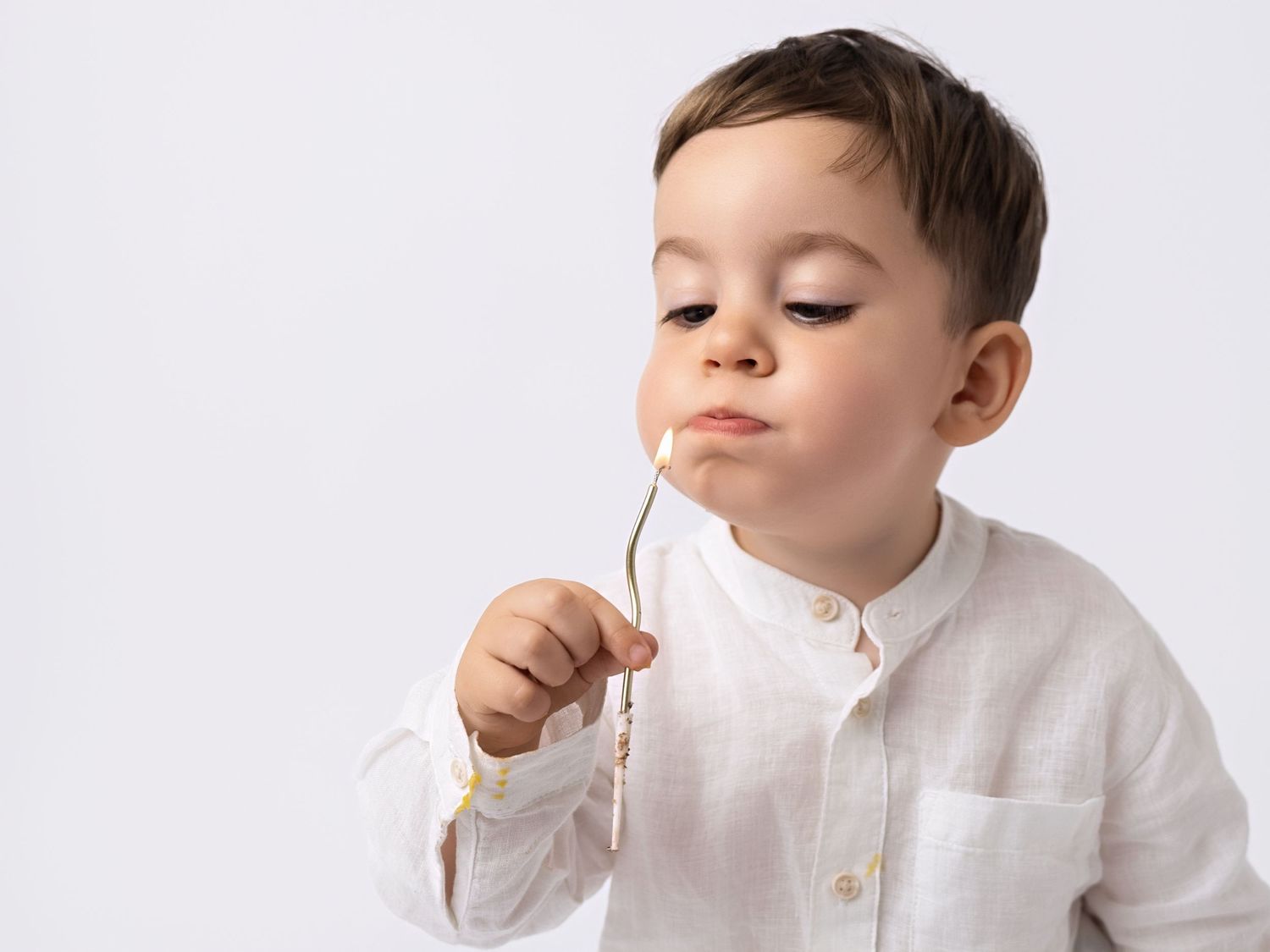 Toddler boy in white linen shirt holding a small flower stem near his mouth on white background.
